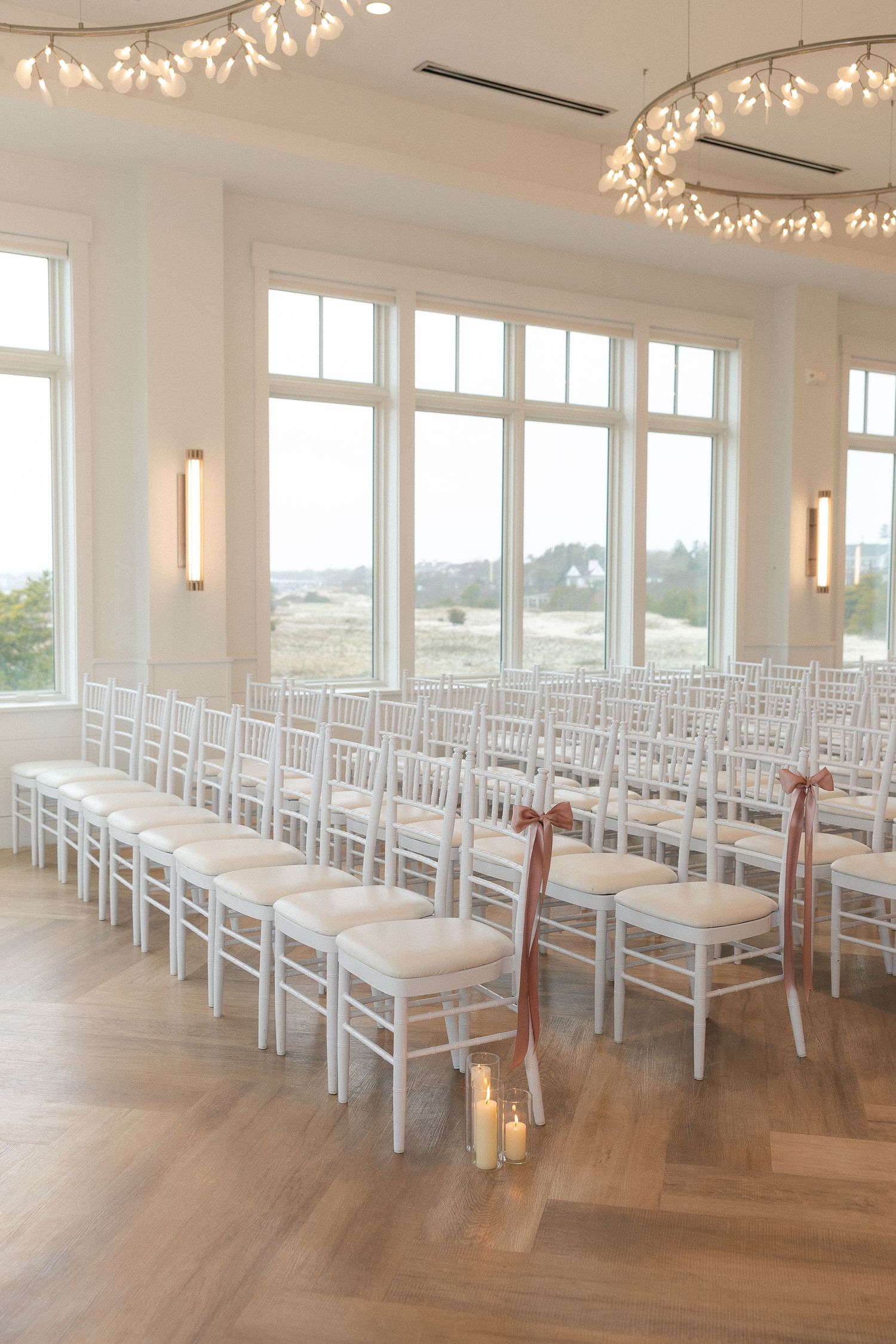 Elegant wedding ceremony venue with white chiavari chairs, herringbone floors, large windows, and warm candlelight accents.