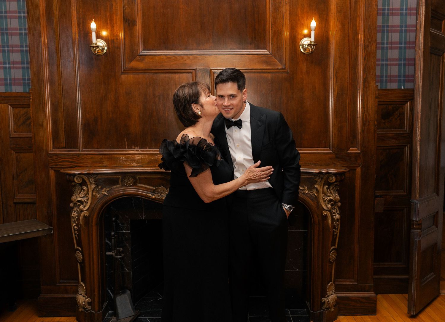Elegant couple in formal attire sharing a romantic moment by a ornate fireplace in a wood-paneled room.