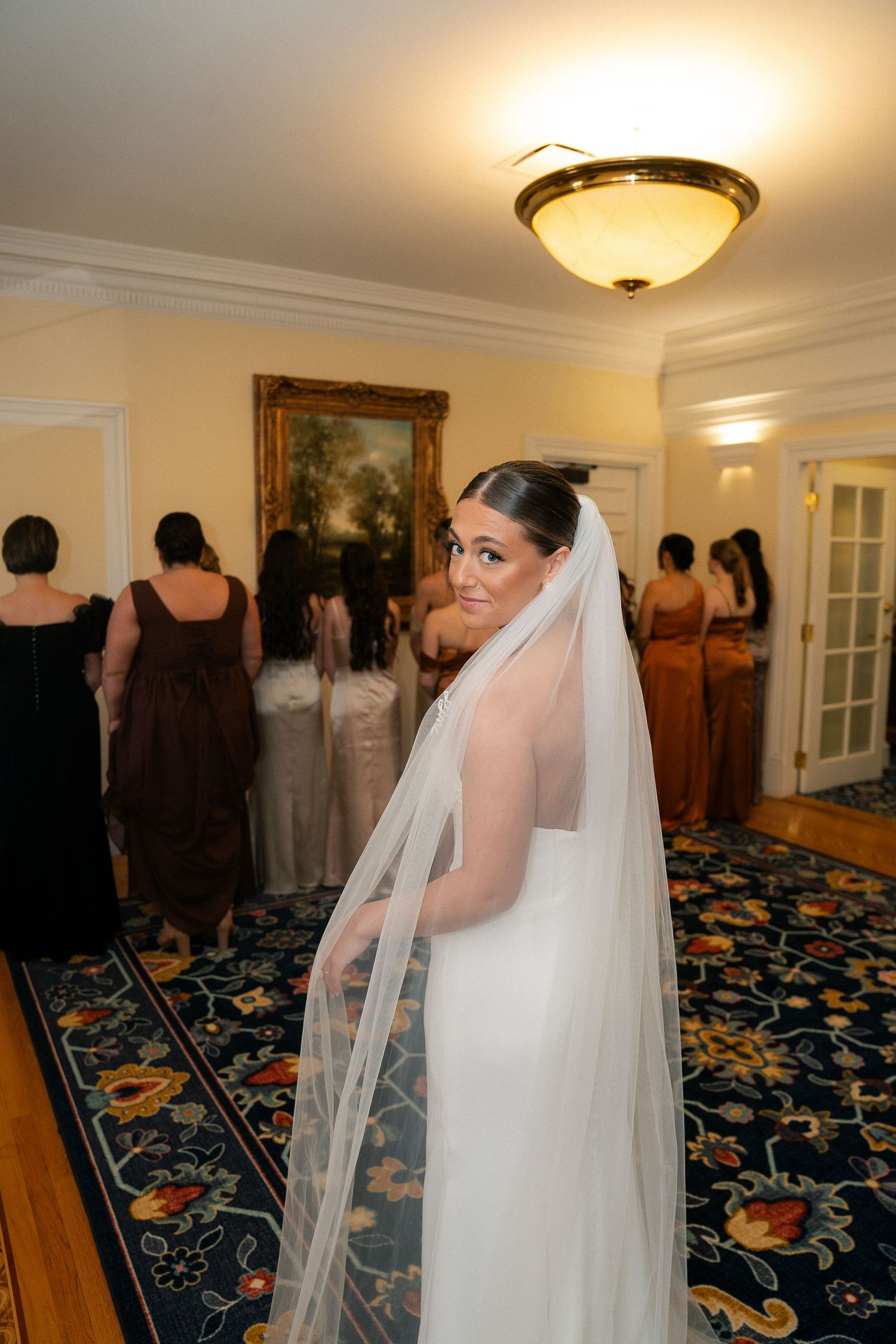 Bride in white gown and long veil smiles over shoulder while bridesmaids gather in elegant room with ornate carpet.