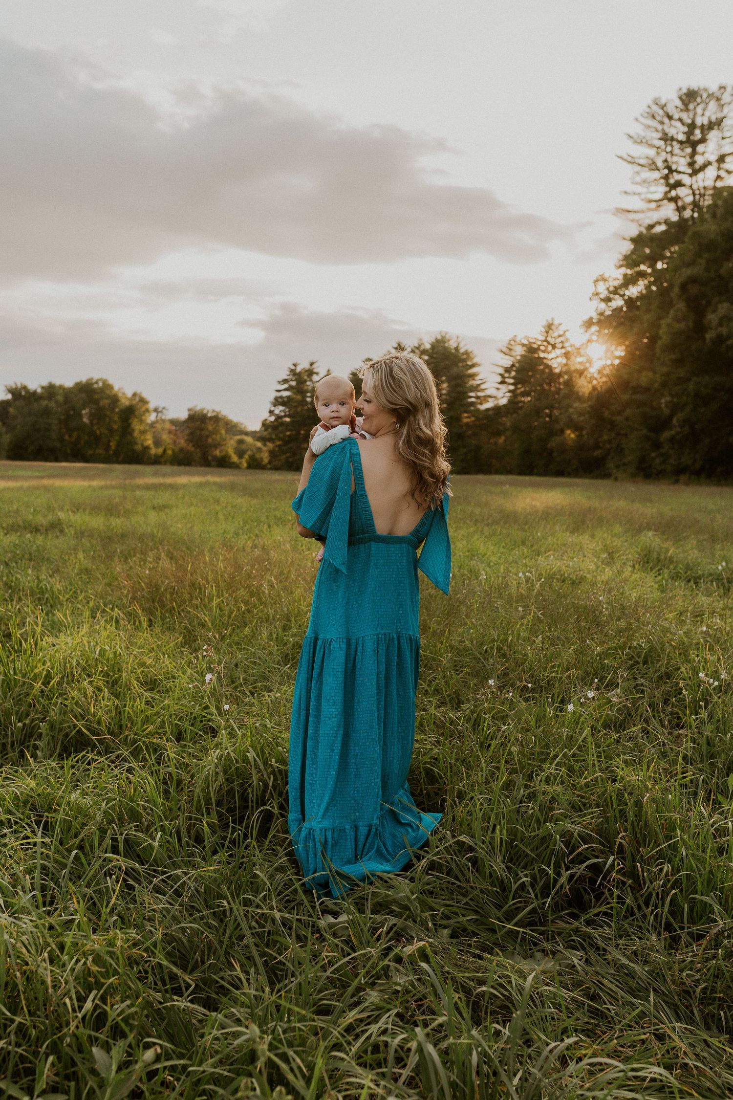 Woman in flowing teal dress stands in golden meadow at sunset holding small child.