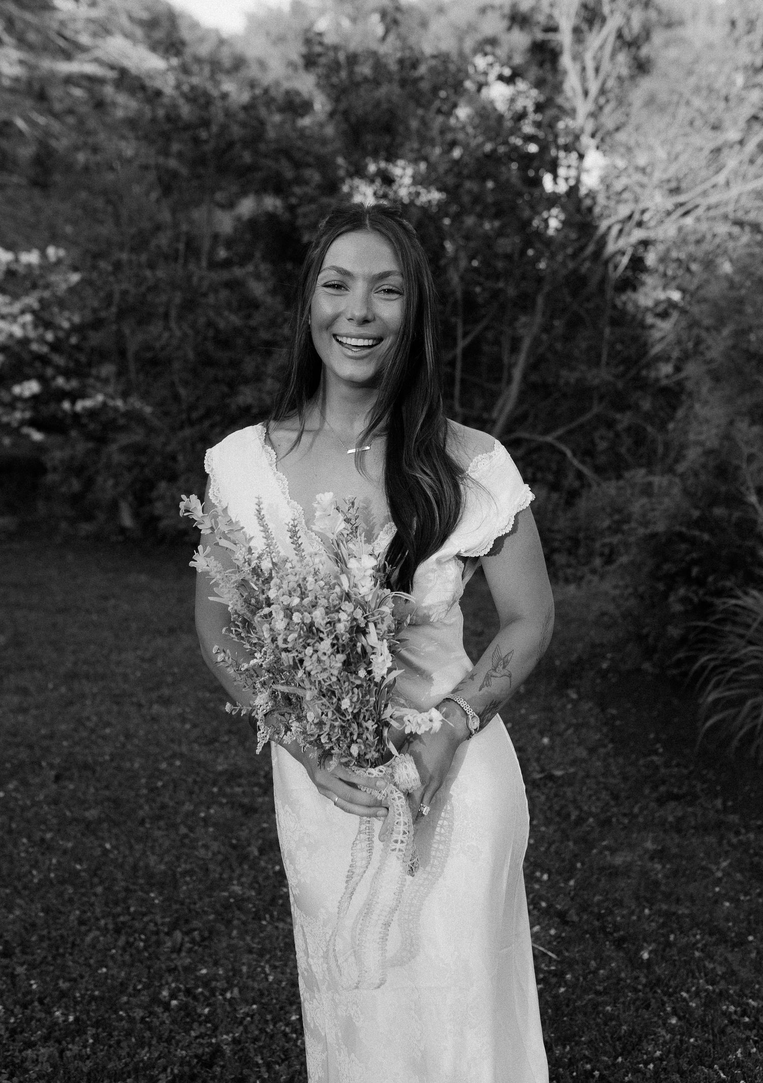Smiling bride in white dress holding wildflower bouquet, standing in lush garden, black and white wedding photo.