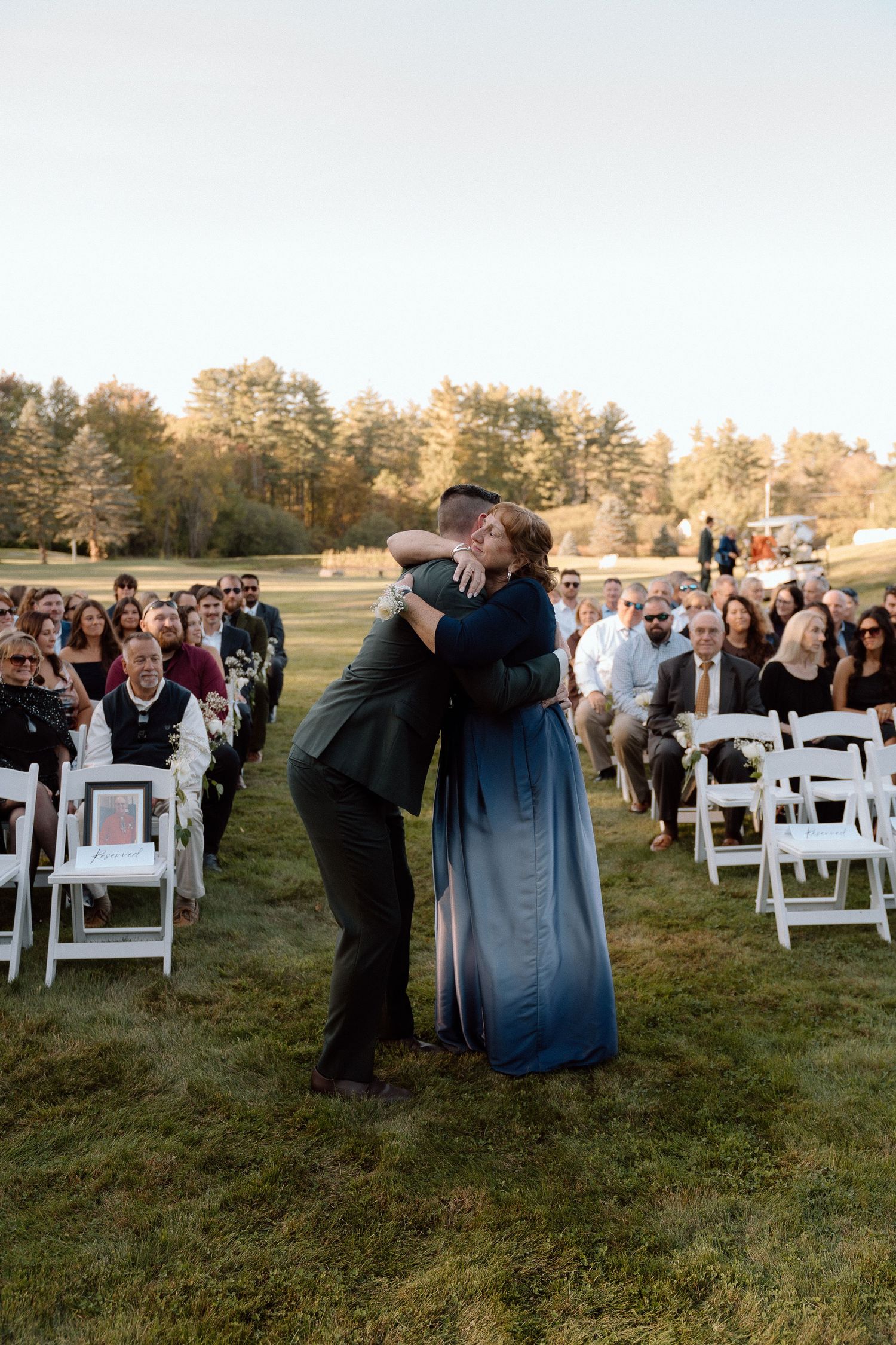 Wedding guests react emotionally during an outdoor ceremony with white chairs on a grassy lawn at sunset.