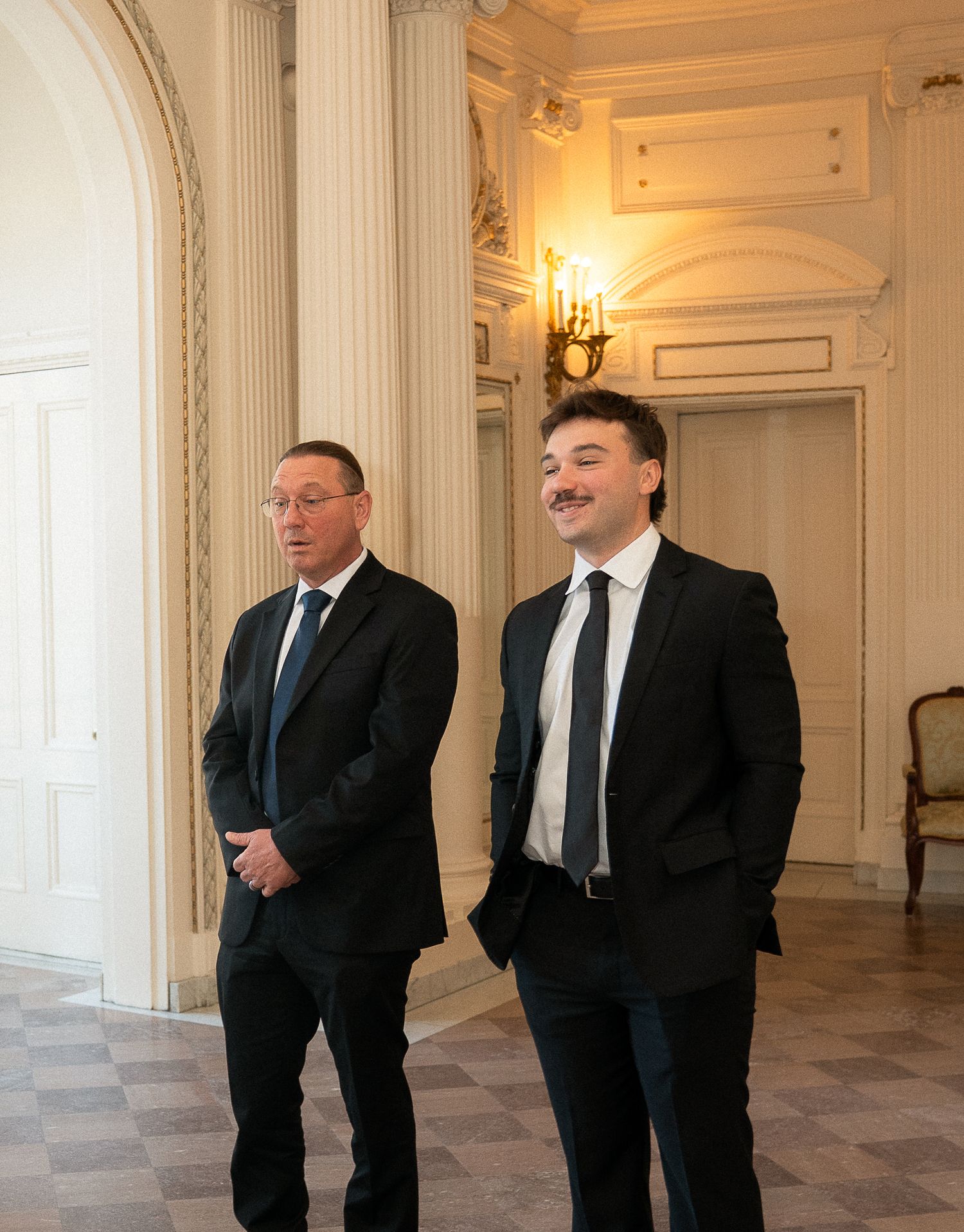 Two men in black suits standing in an elegant room with white columns and ornate wall sconces.