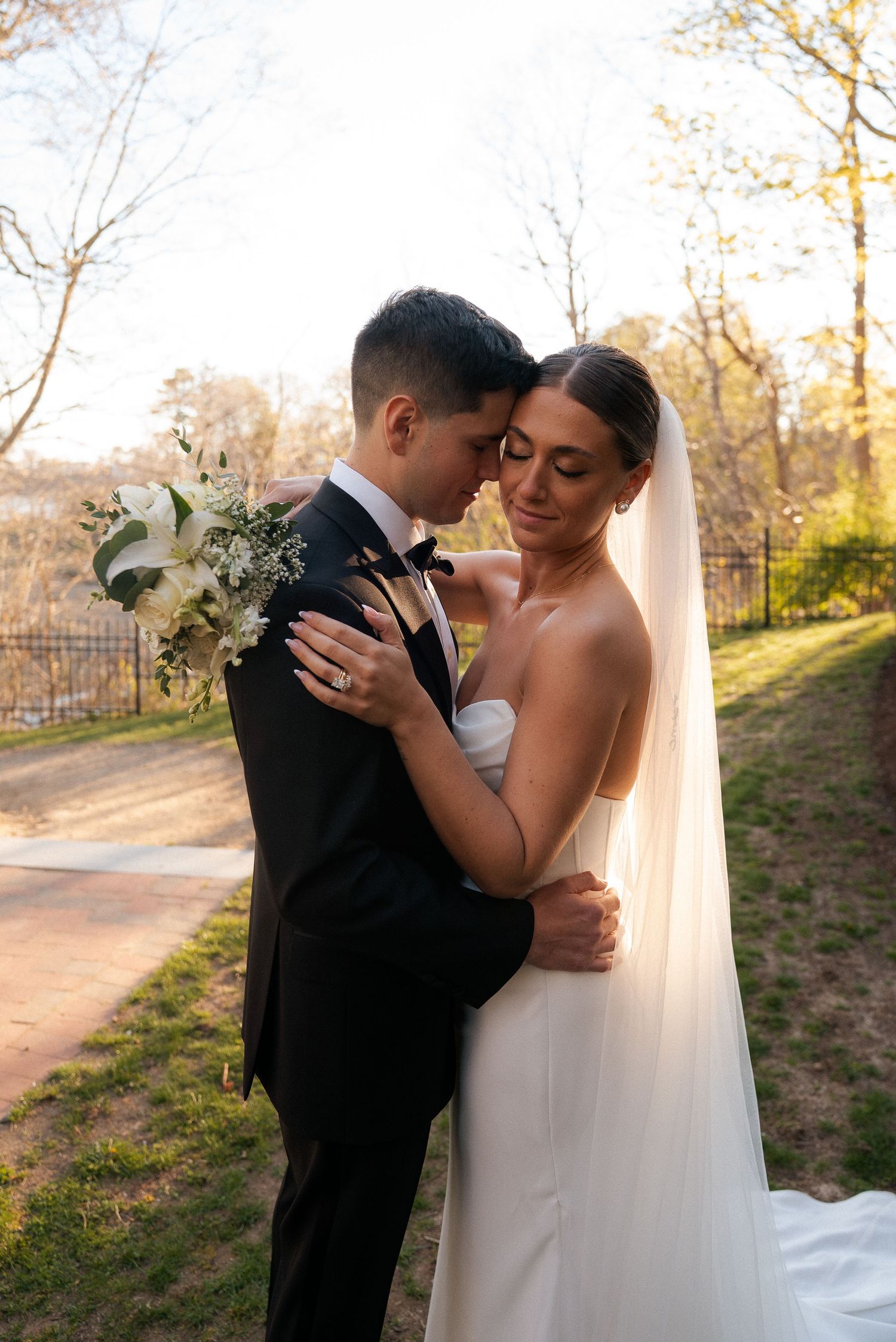 Bride and groom embrace tenderly in golden sunset light with white floral bouquet outdoors.