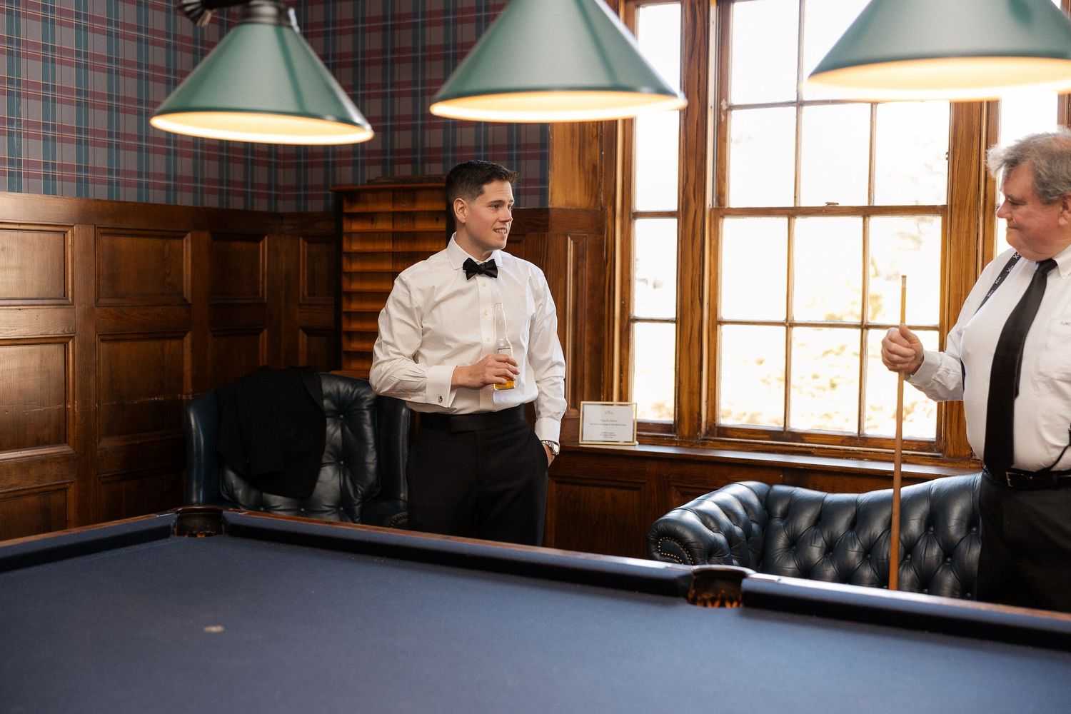 Two men in formal attire socializing near a pool table in an elegant wood-paneled billiards room.