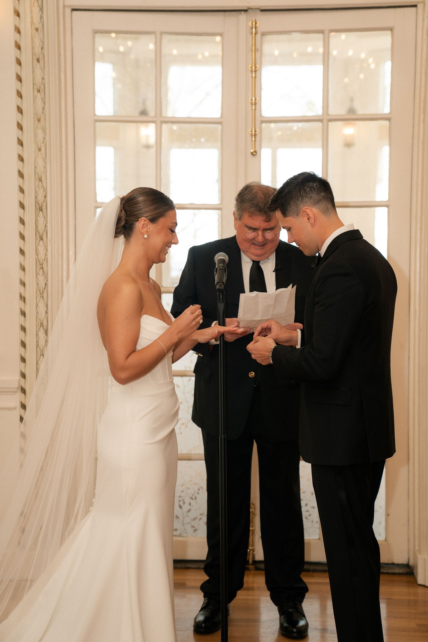 Smiling bride places wedding ring on groom's finger as officiant watches during indoor ceremony.
