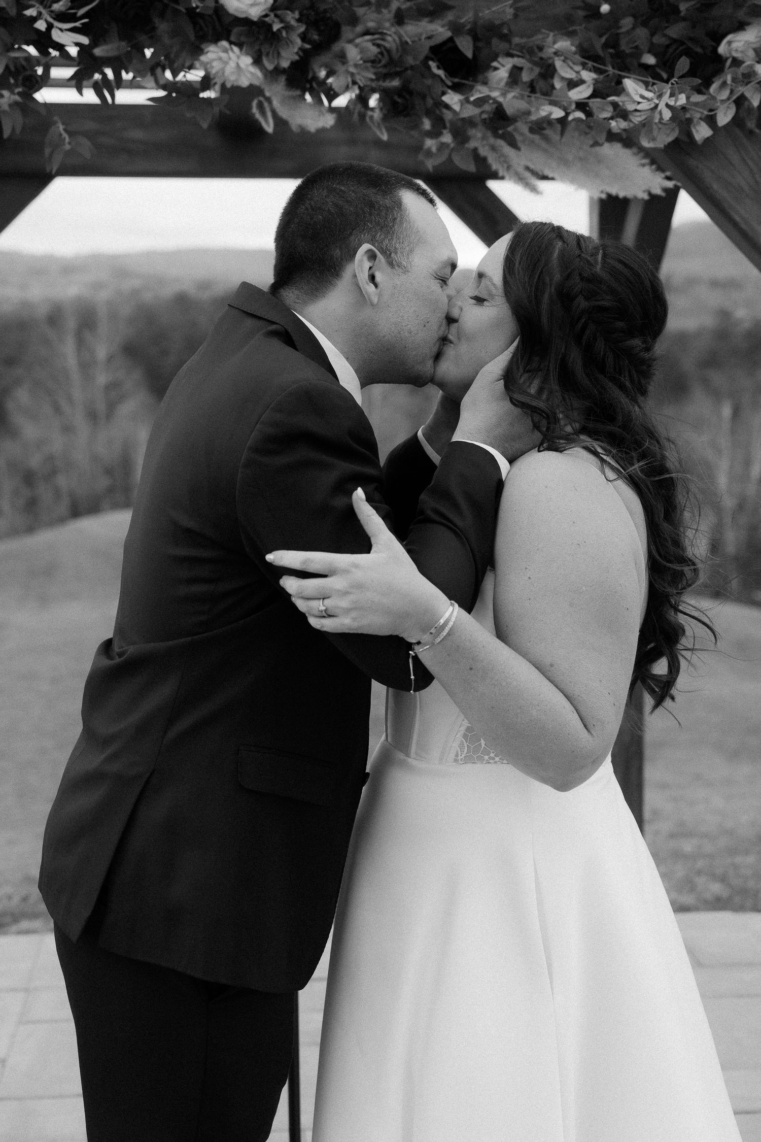 A romantic wedding couple shares a tender kiss outdoors in formal black and white attire.