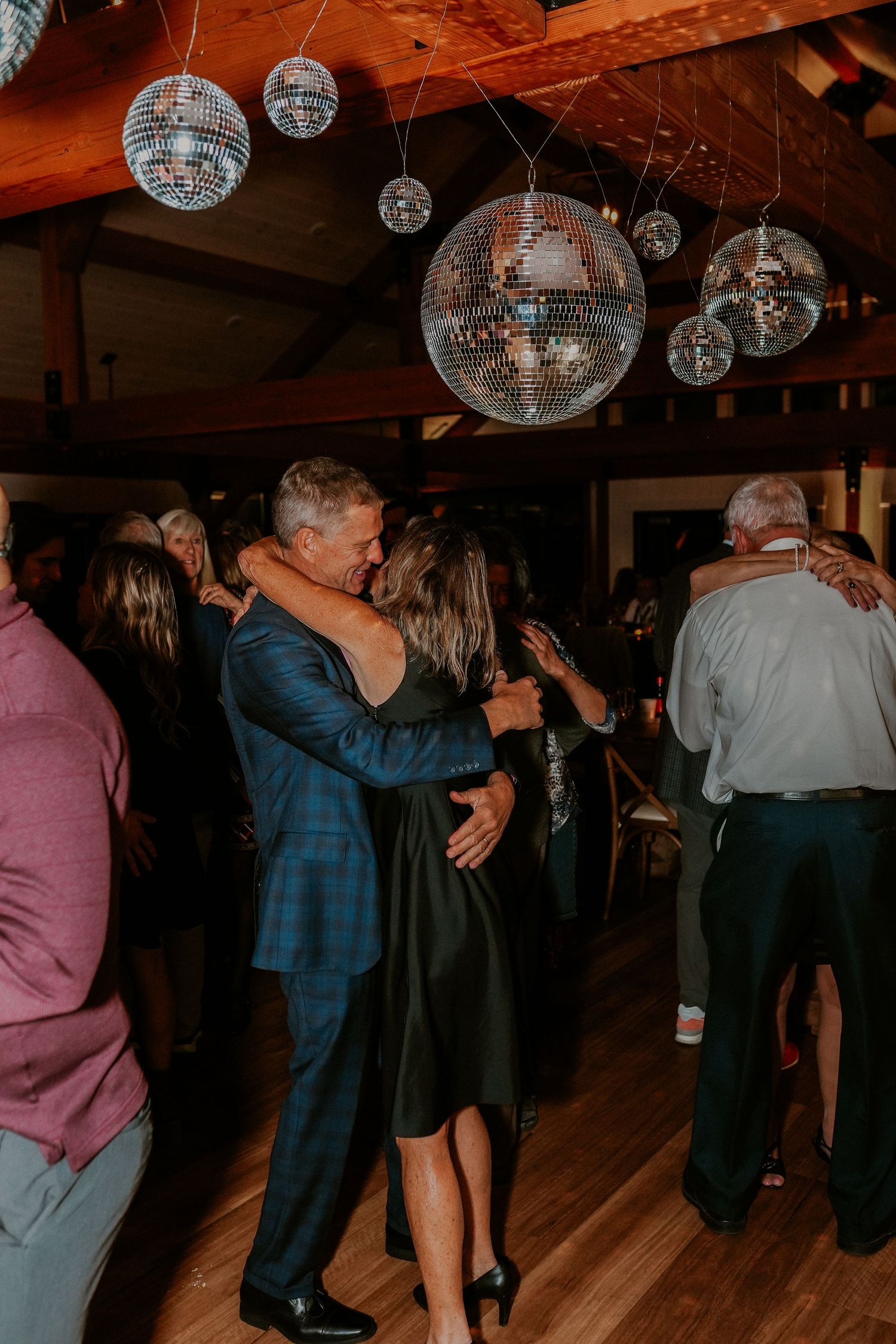 Couples dancing together under disco balls at a dimly lit venue with wooden floors.