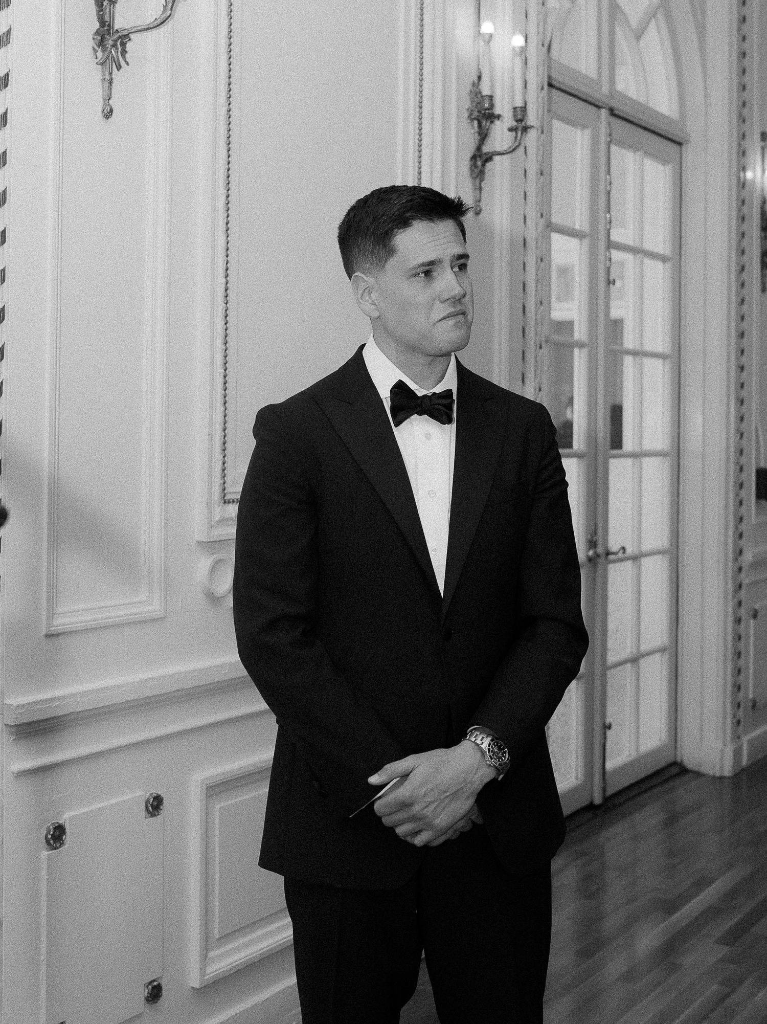 A groom in a black tuxedo and bow tie stands elegantly in a grand white ballroom, hands clasped.