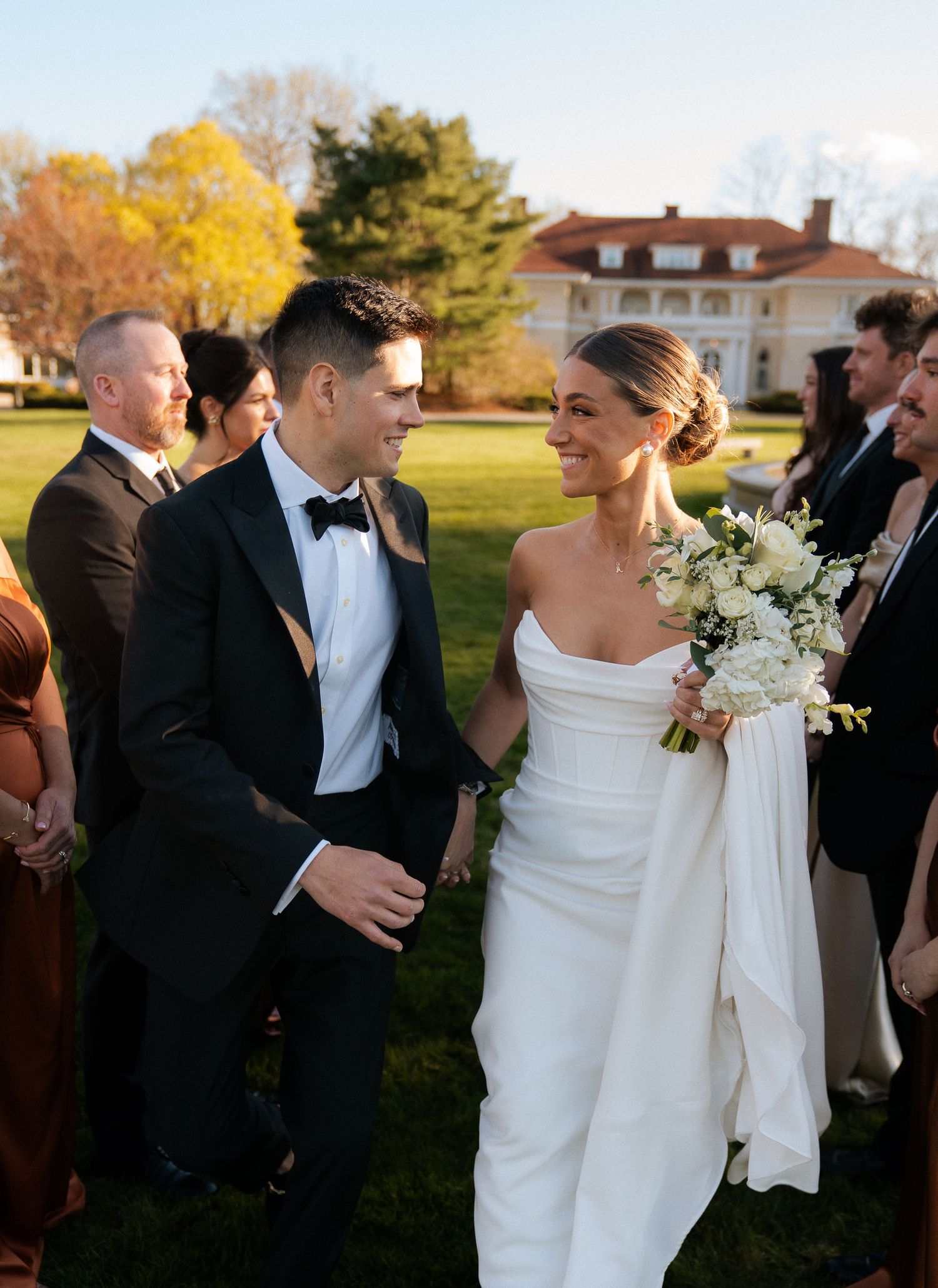 Bride in strapless white gown holding white bouquet walks with tuxedo-clad groom past guests at outdoor wedding ceremony.