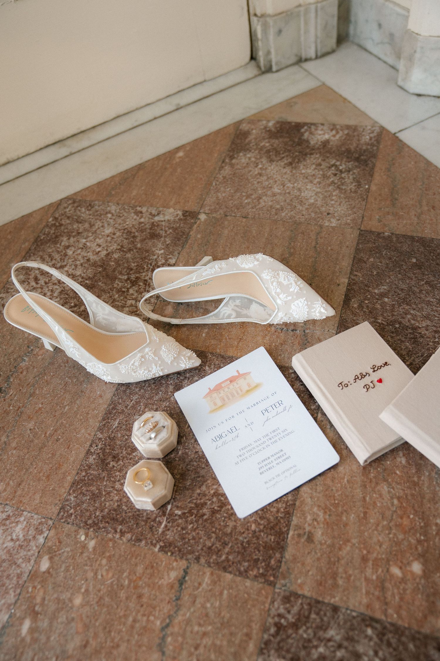 Elegant wedding flat lay with lace bridal heels, ring boxes, invitation suite, and a handwritten note on stone floor.