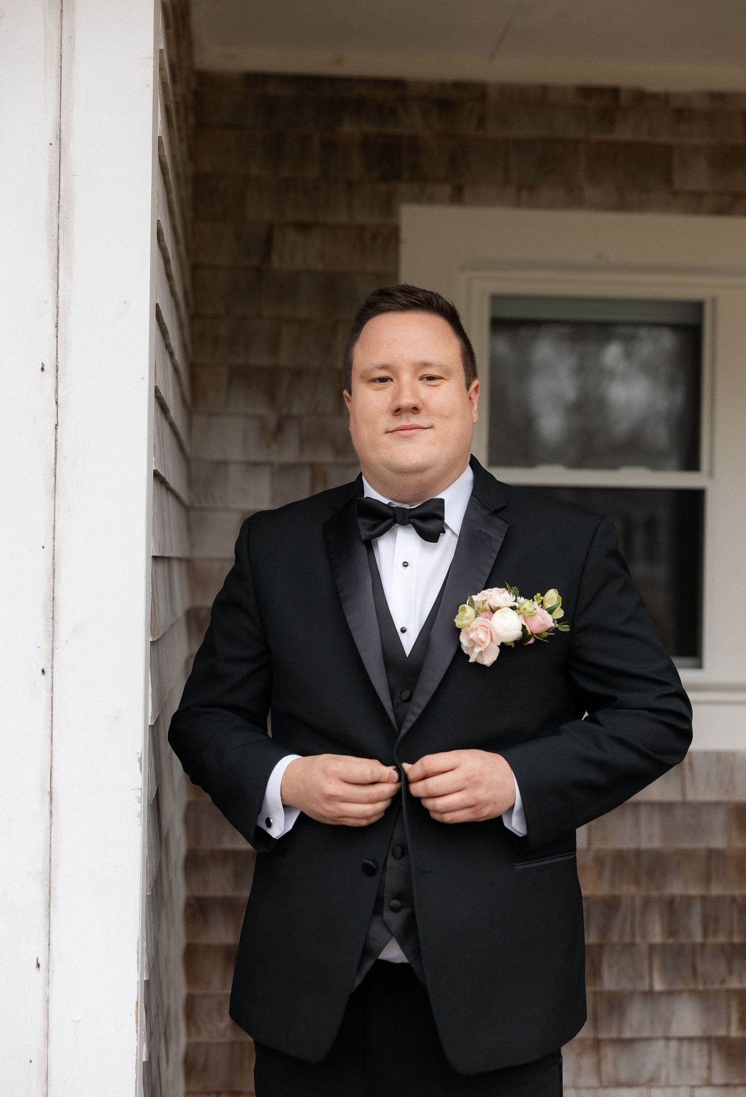 Groom in black tuxedo with bow tie and floral boutonniere buttons jacket outside a shingle-style home.