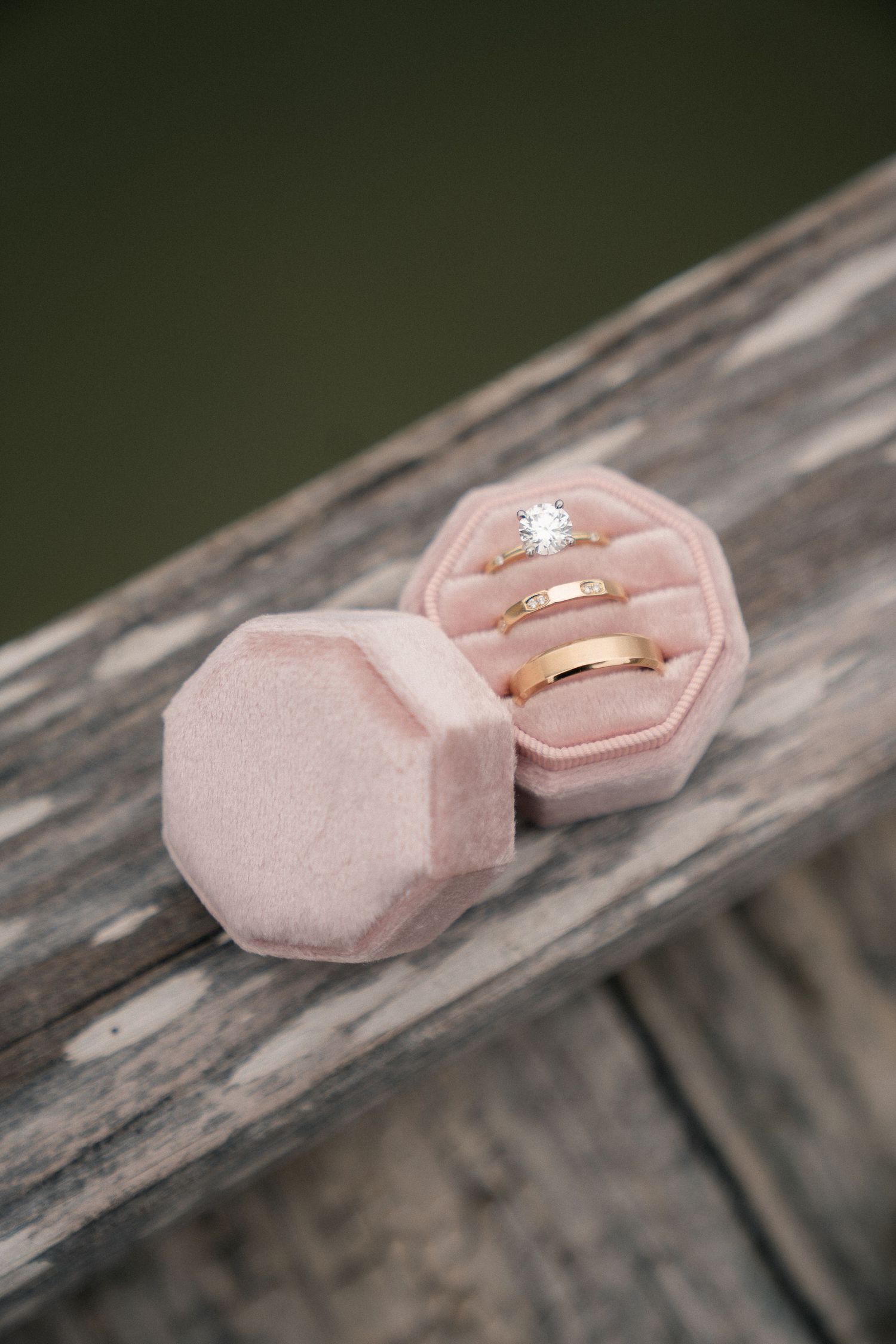 Pink velvet octagonal ring box holding a diamond engagement ring and two gold wedding bands on rustic wood.