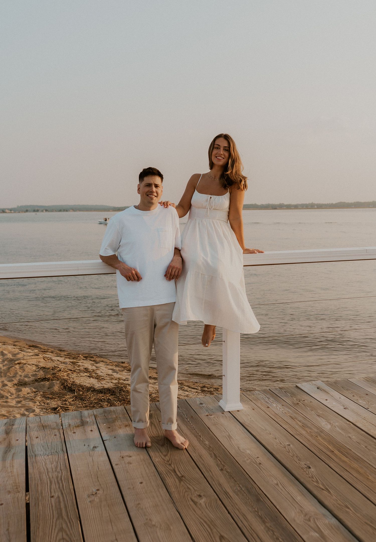 A couple in coordinated white outfits pose together on a wooden dock by the water.