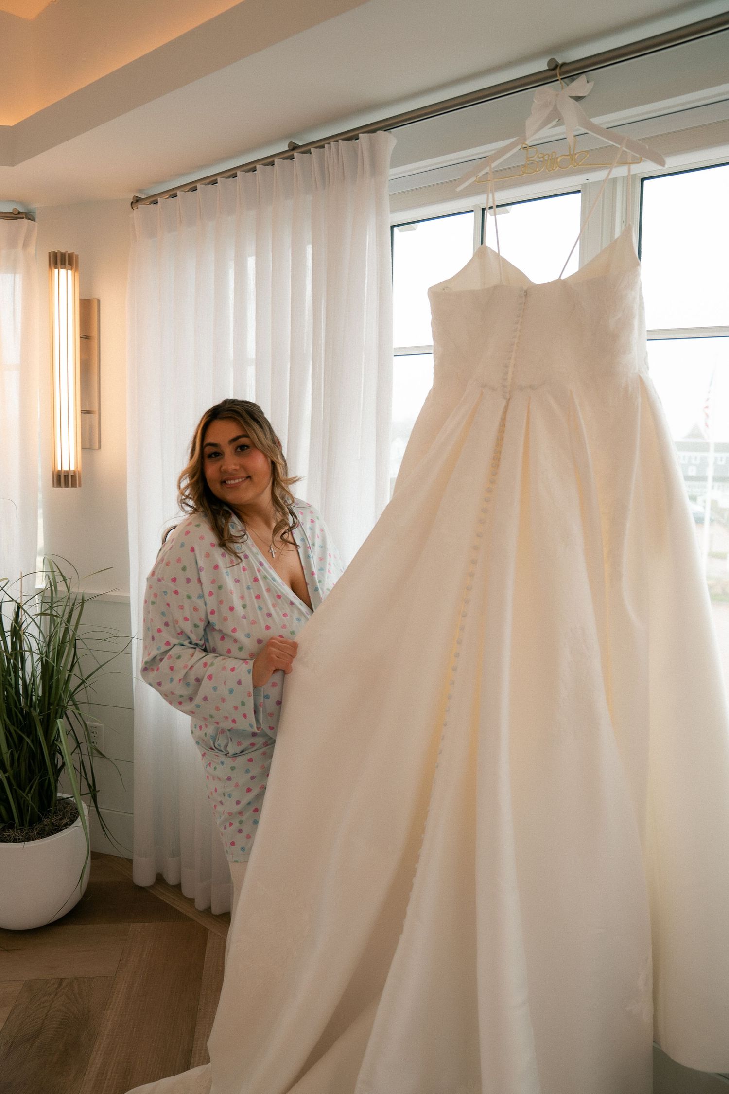 Smiling bride in floral pajamas standing beside her elegant strapless ball gown wedding dress.