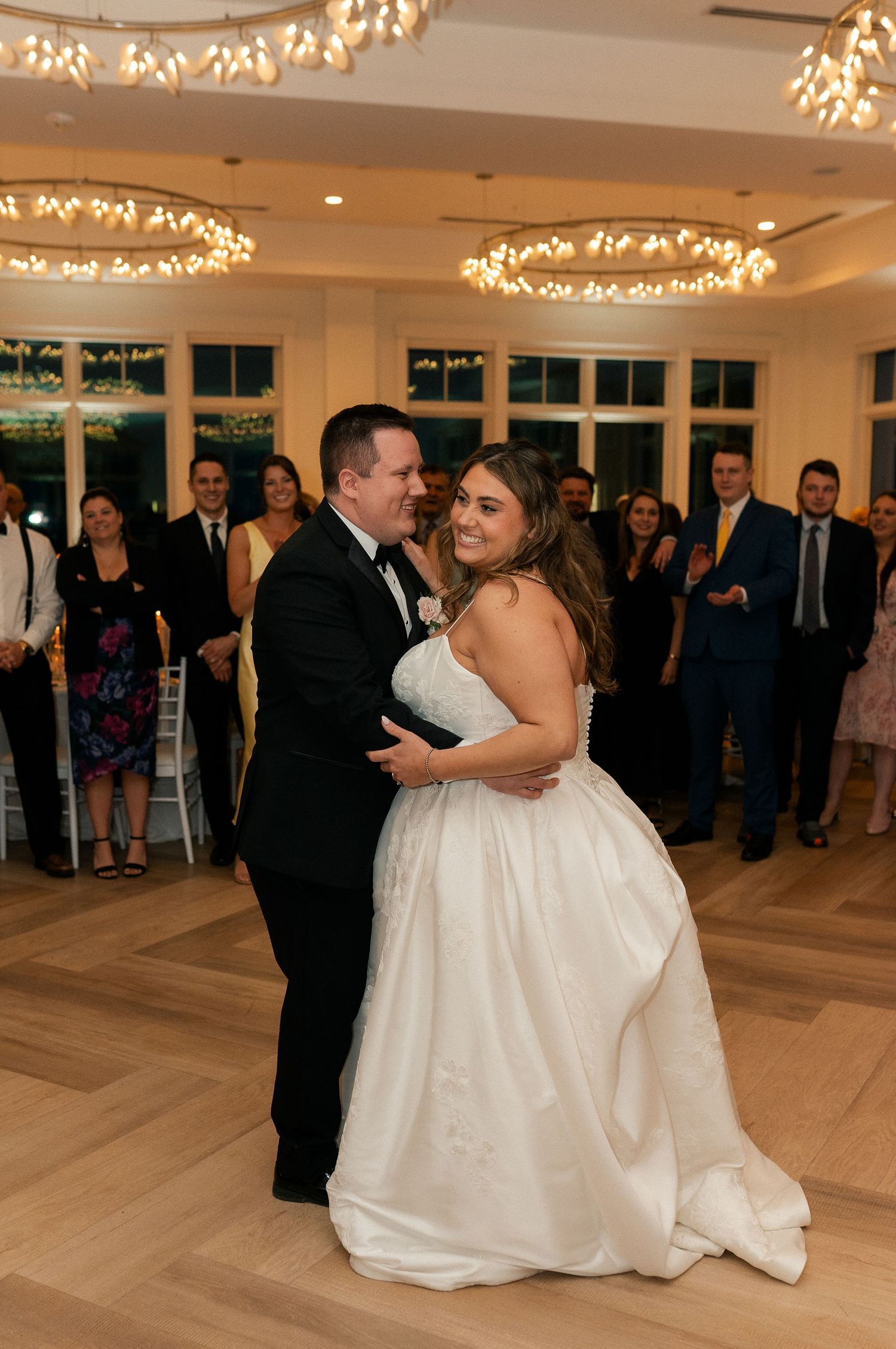 Bride and groom sharing their first dance surrounded by applauding guests in a warmly lit ballroom.