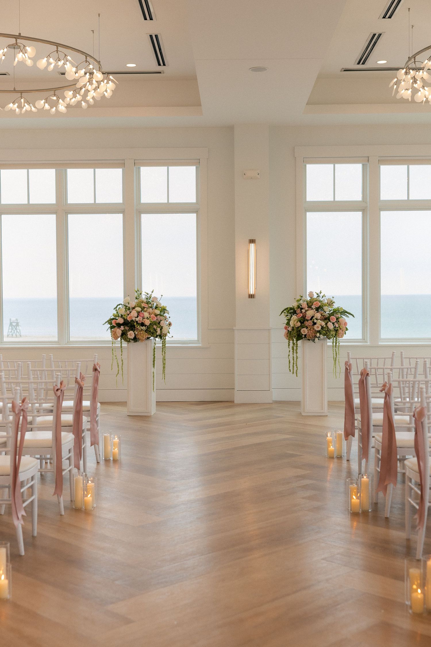 Elegant indoor wedding ceremony setup with floral arrangements, candles, and ocean views through large white-framed windows.