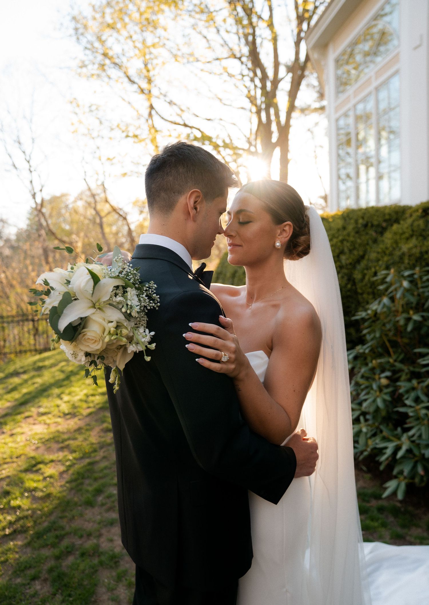 Couple shares intimate moment in glowing backlit sunlight with white bridal bouquet and veil.