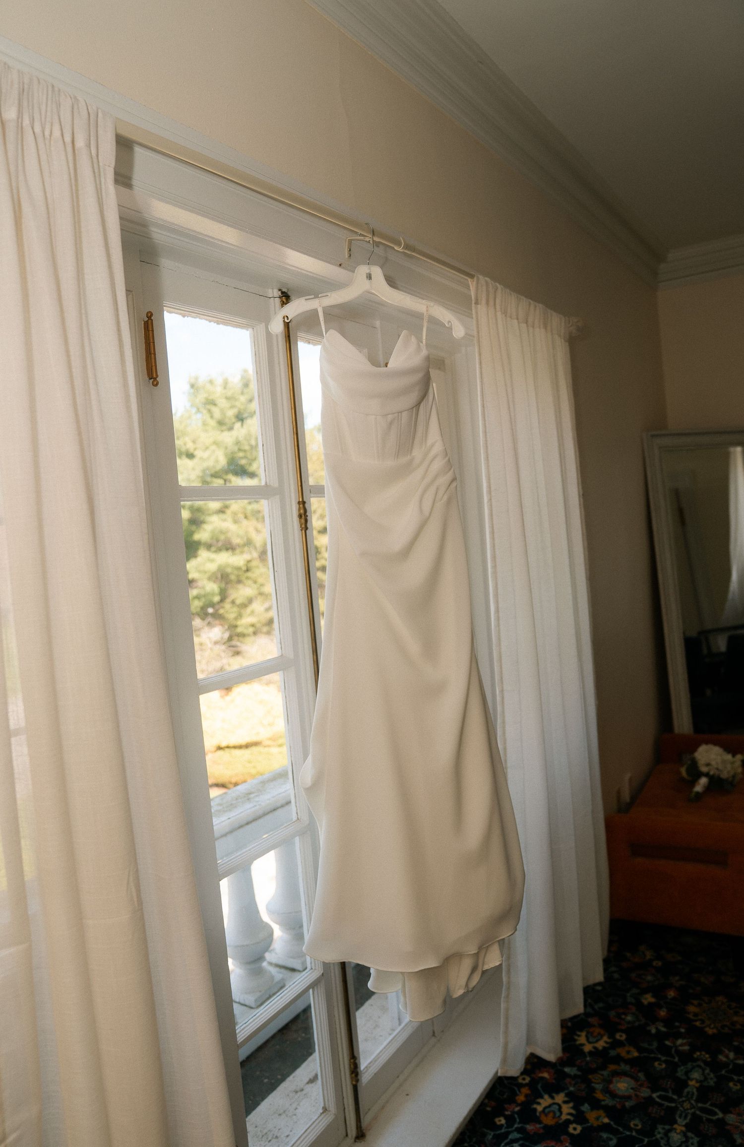 Elegant white strapless wedding gown hanging on a curtain rod in front of a sunlit window.