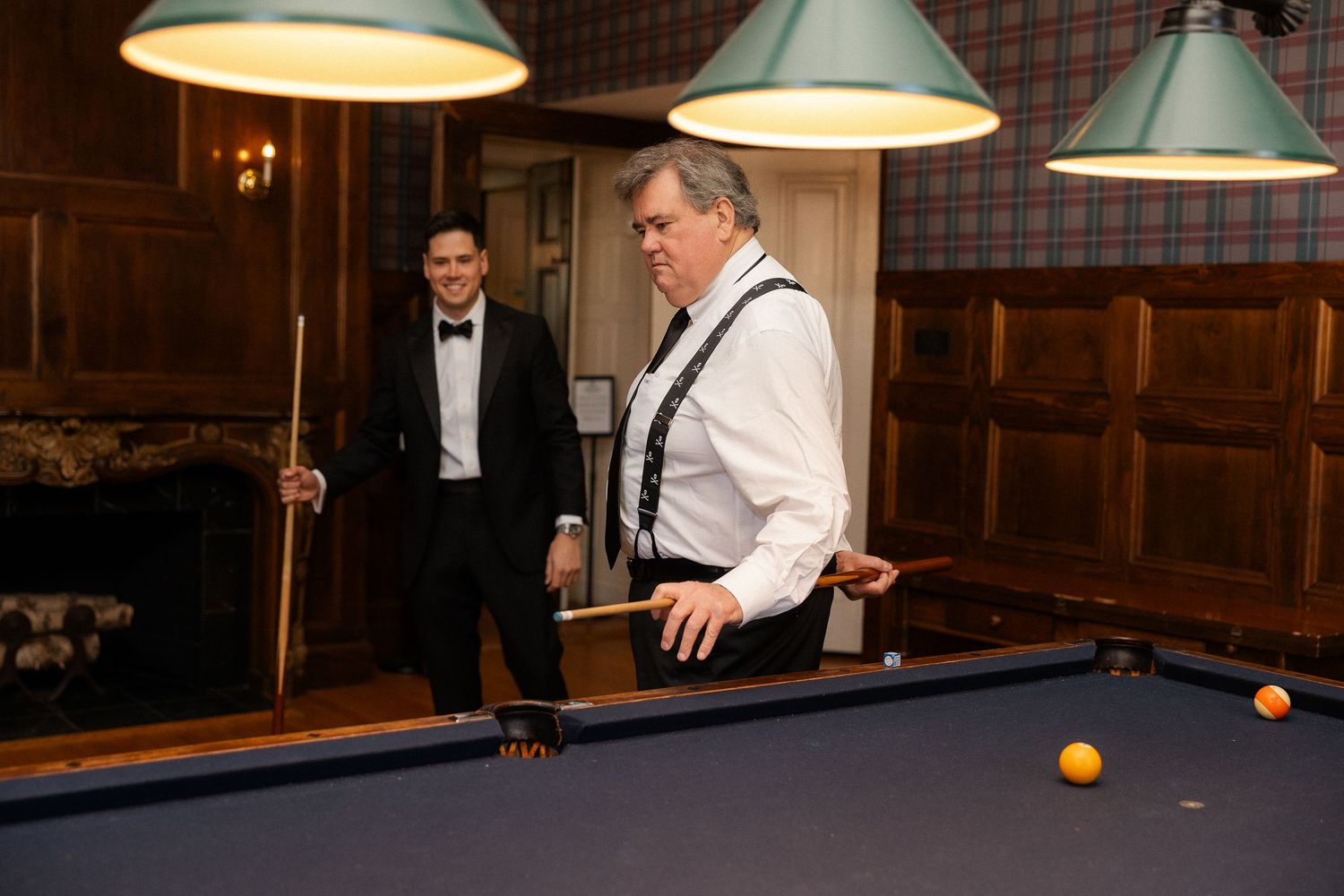 Two men in formal attire playing billiards in an elegant wood-paneled room with green pendant lights.