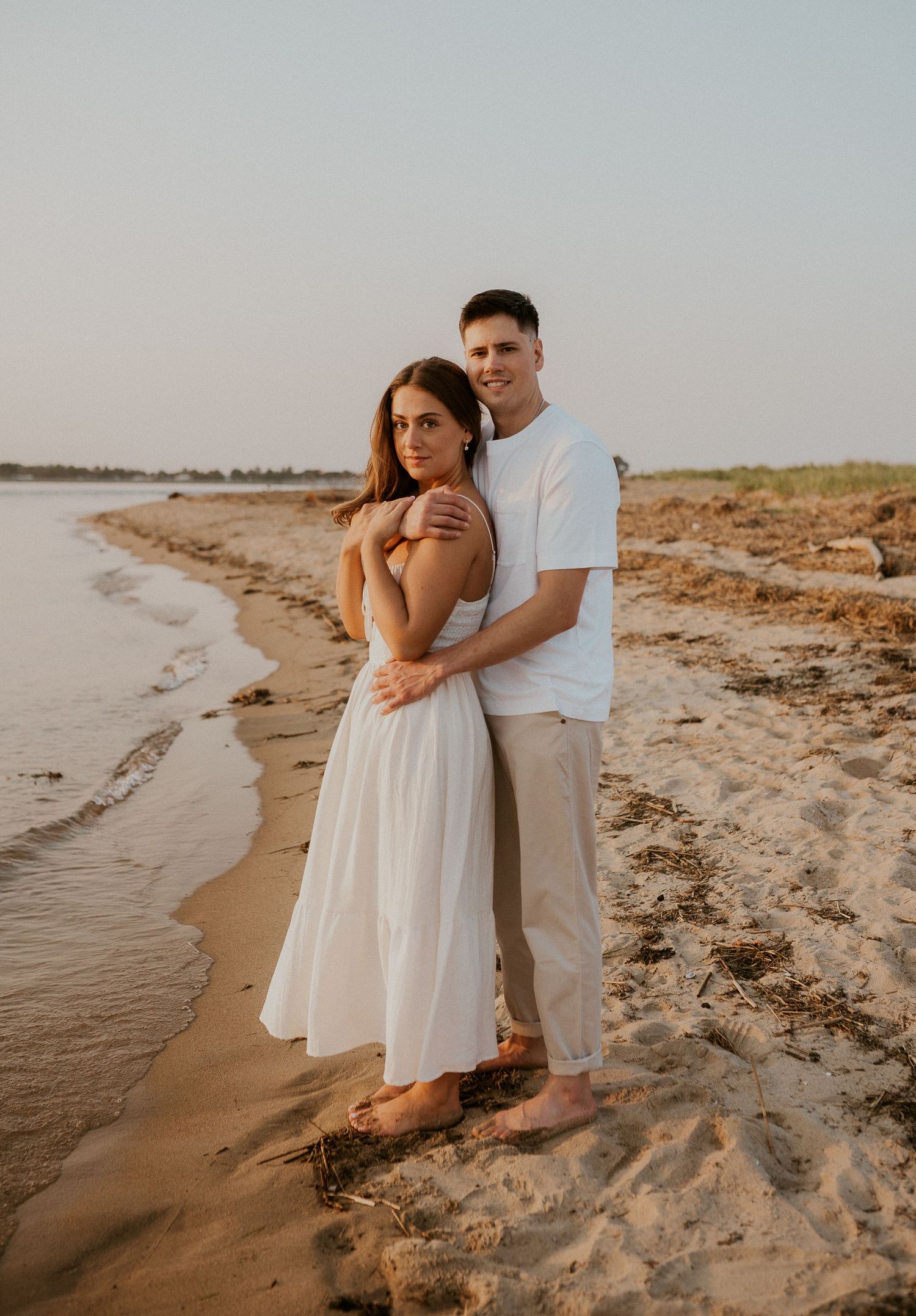 Couple shares tender moments on a sandy beach wearing white summer clothing at dusk.
