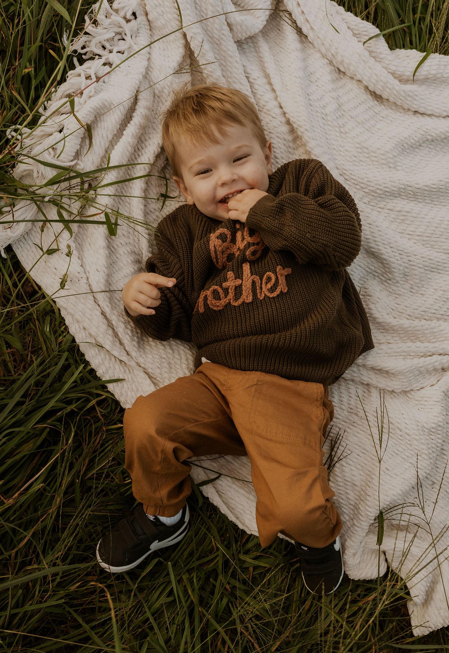 Toddler in brown sweater smiling while lying on white blanket in grass.