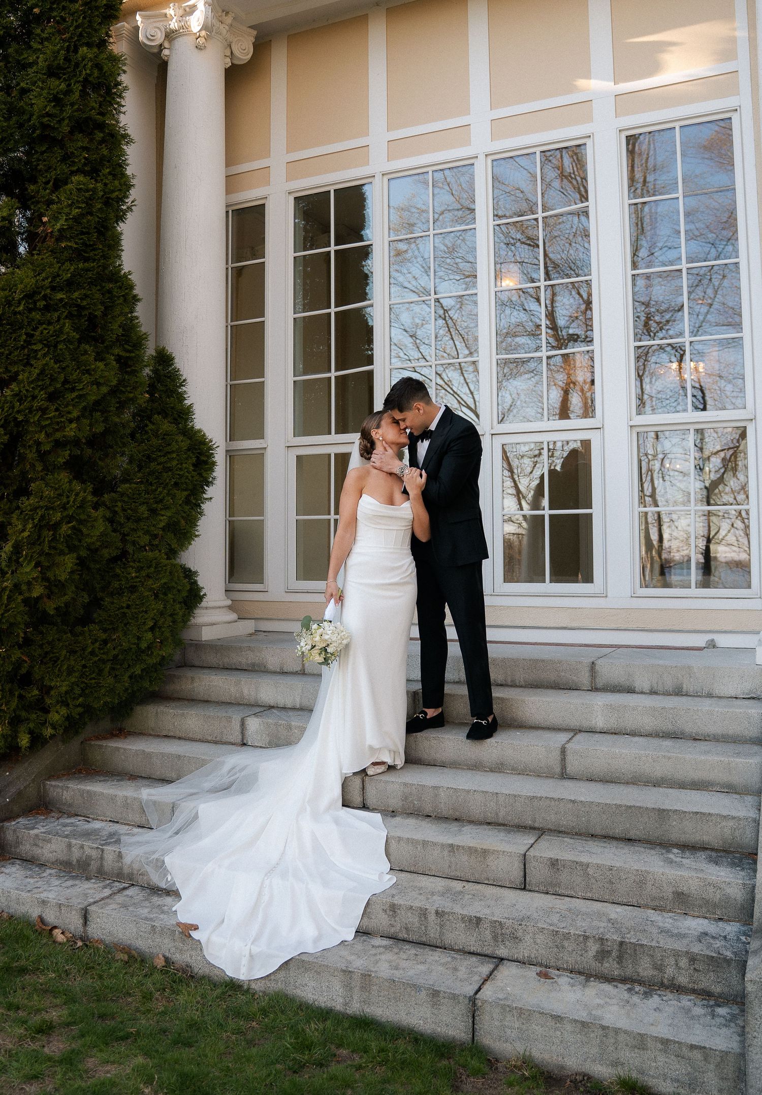 Bride and groom share a romantic moment on stone steps outside an elegant venue with tall windows.