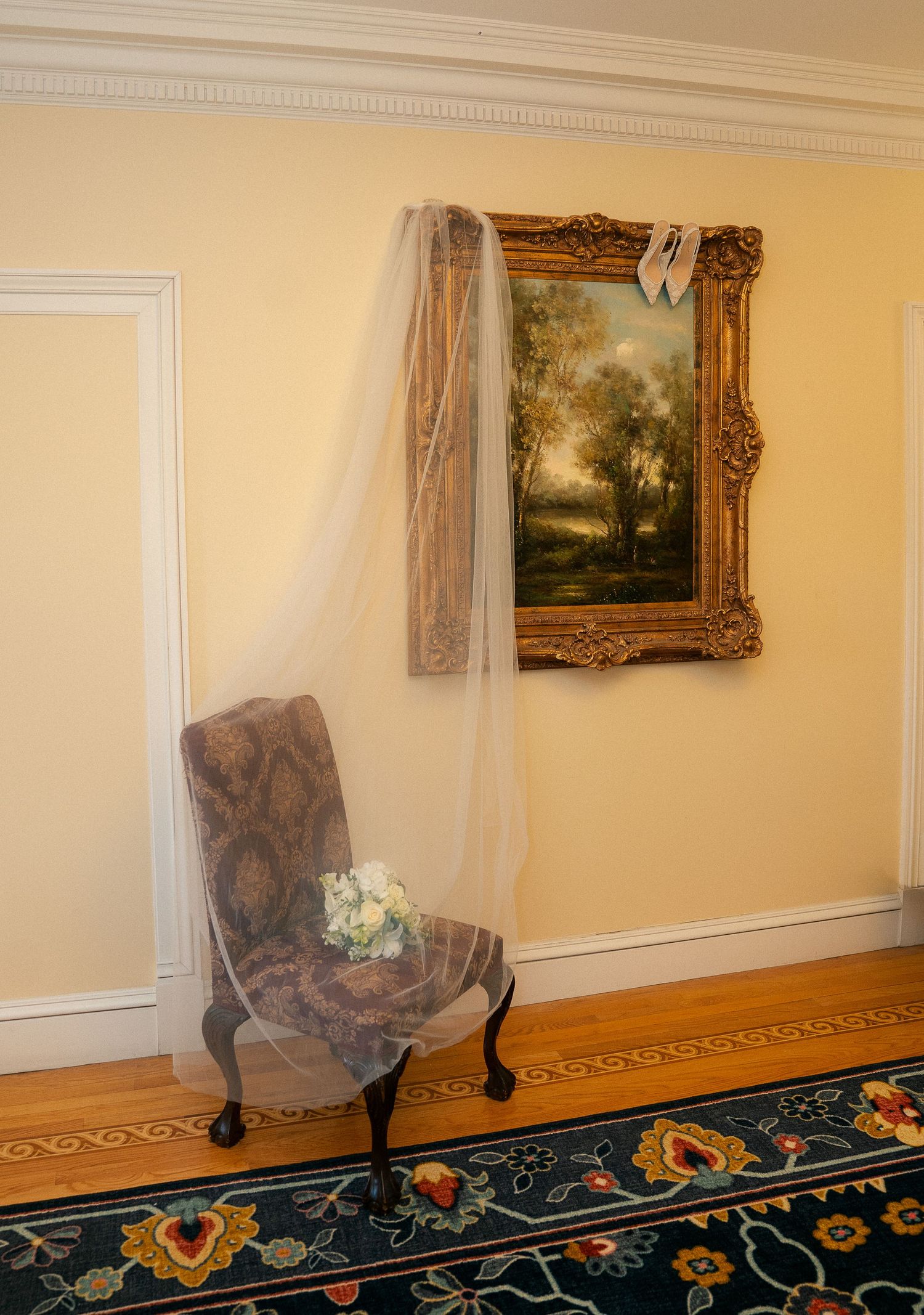 Elegant wedding veil draped over ornate gold-framed landscape painting above a vintage chair with bridal bouquet.