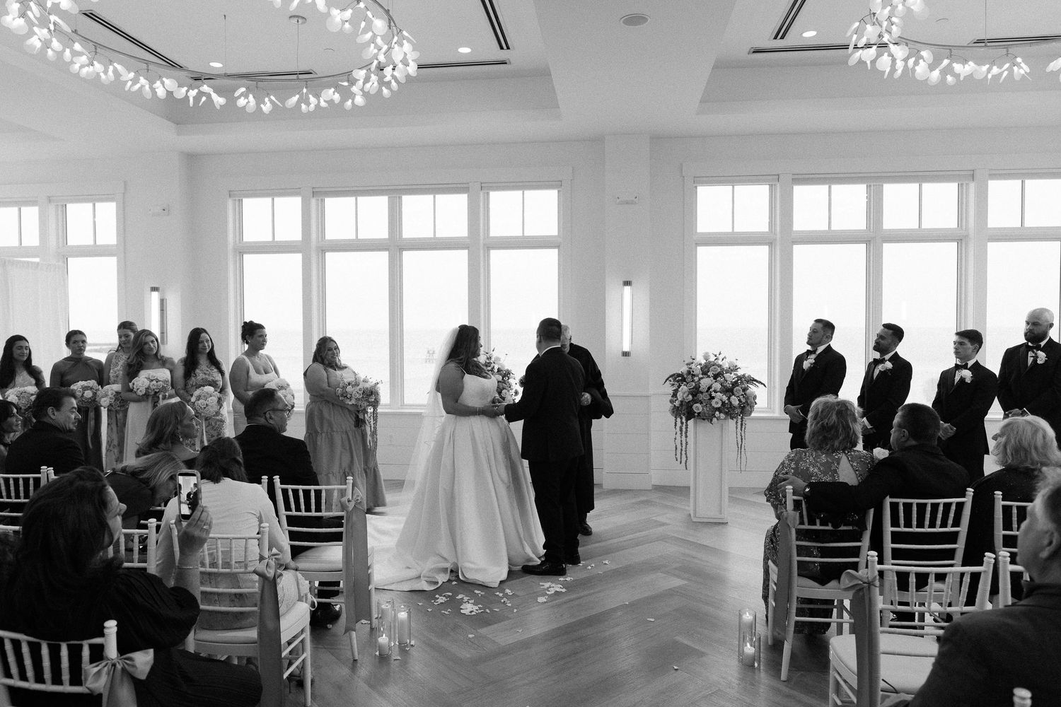 Black and white photo of a wedding ceremony in an elegant venue with large windows, guests seated as couple exchanges vows.
