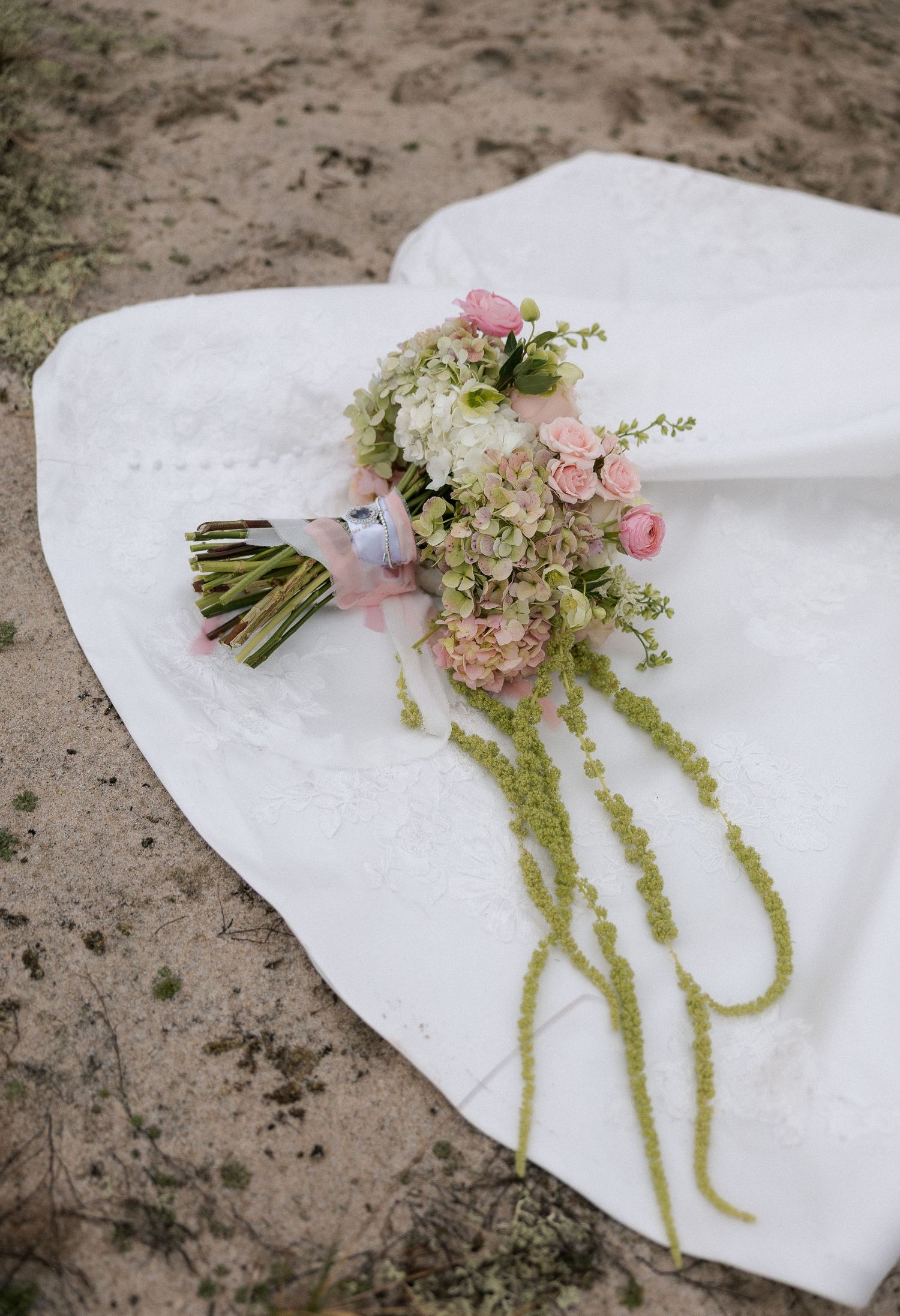Bridal bouquet of pink roses and hydrangeas with green trailing accents on white wedding dress.