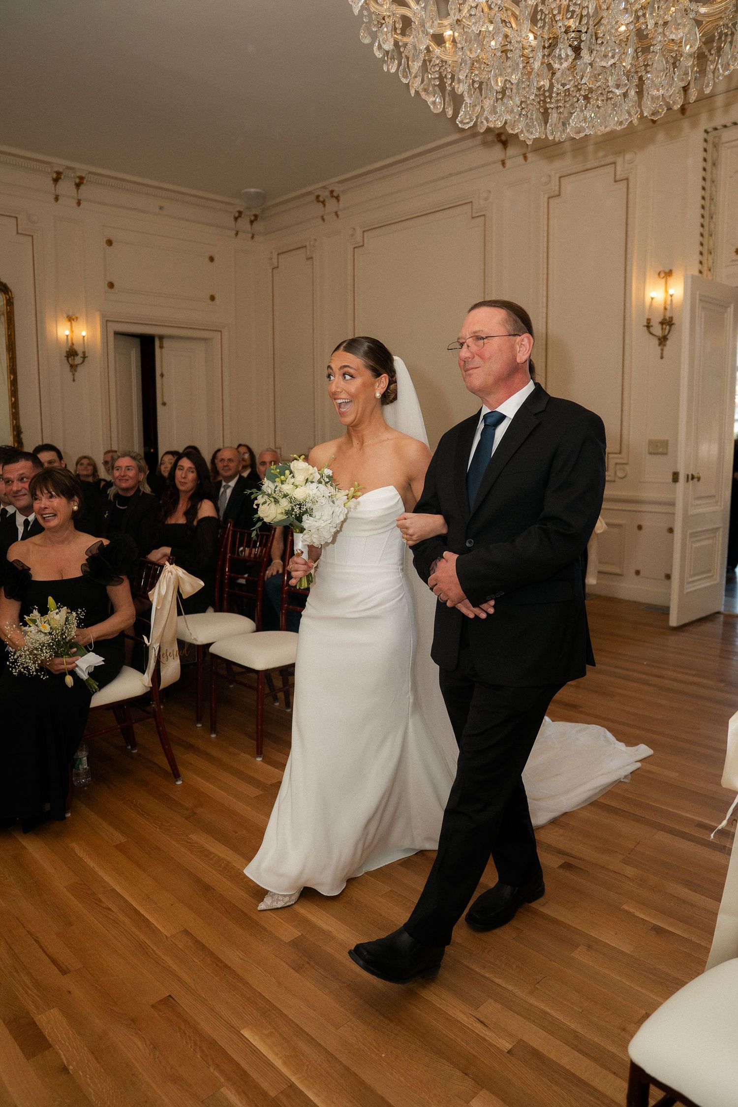 Bride in white gown walks down aisle with father in elegant ballroom during wedding ceremony.