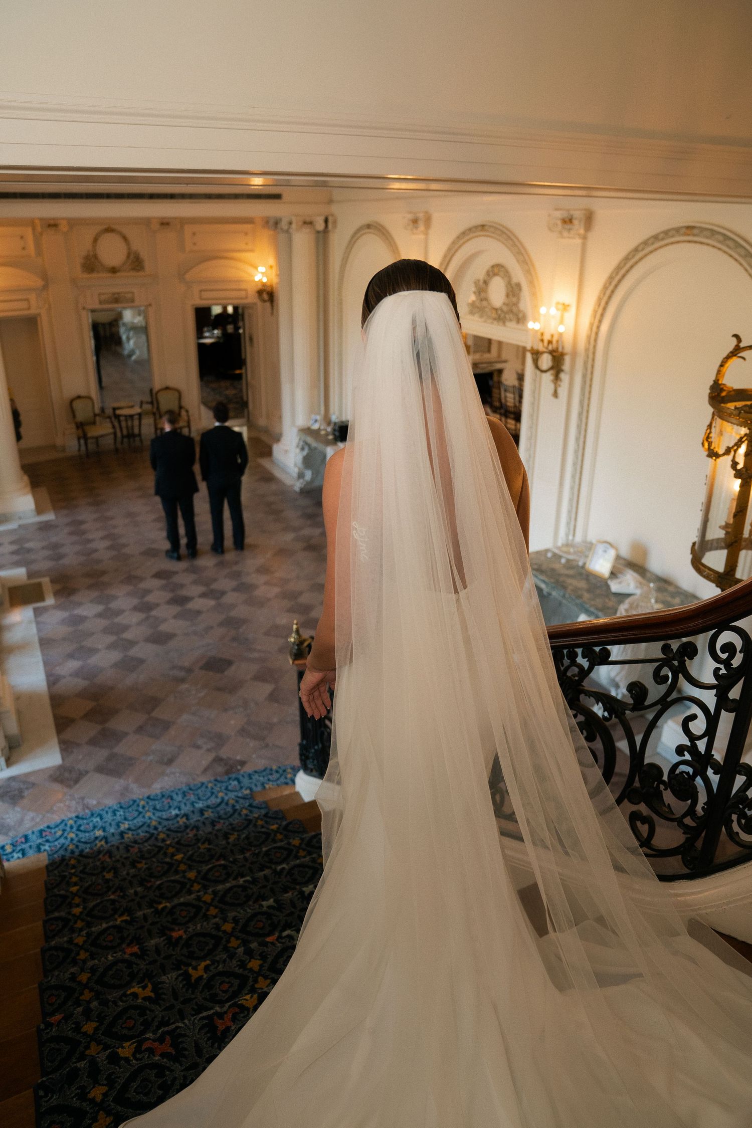 Bride in elegant white wedding gown and long veil stands at top of grand staircase in ornate venue.