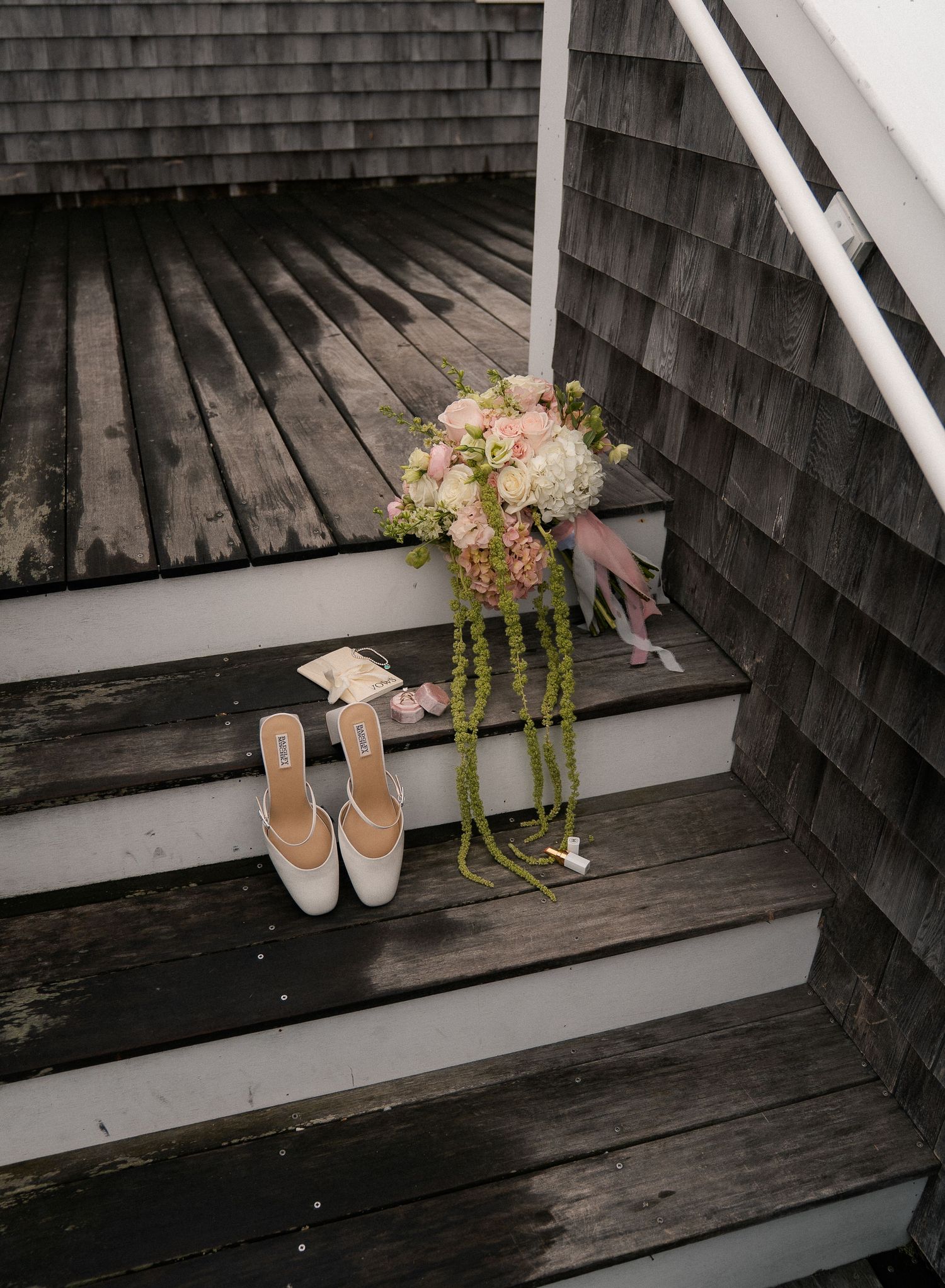 Bridal heels and pink floral bouquet with trailing greenery displayed on rustic wooden steps of a coastal home.