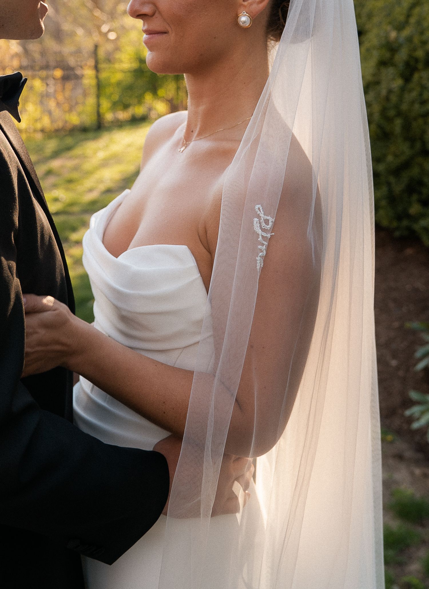 Bride in strapless white gown with embroidered veil embracing groom in black suit outdoors.