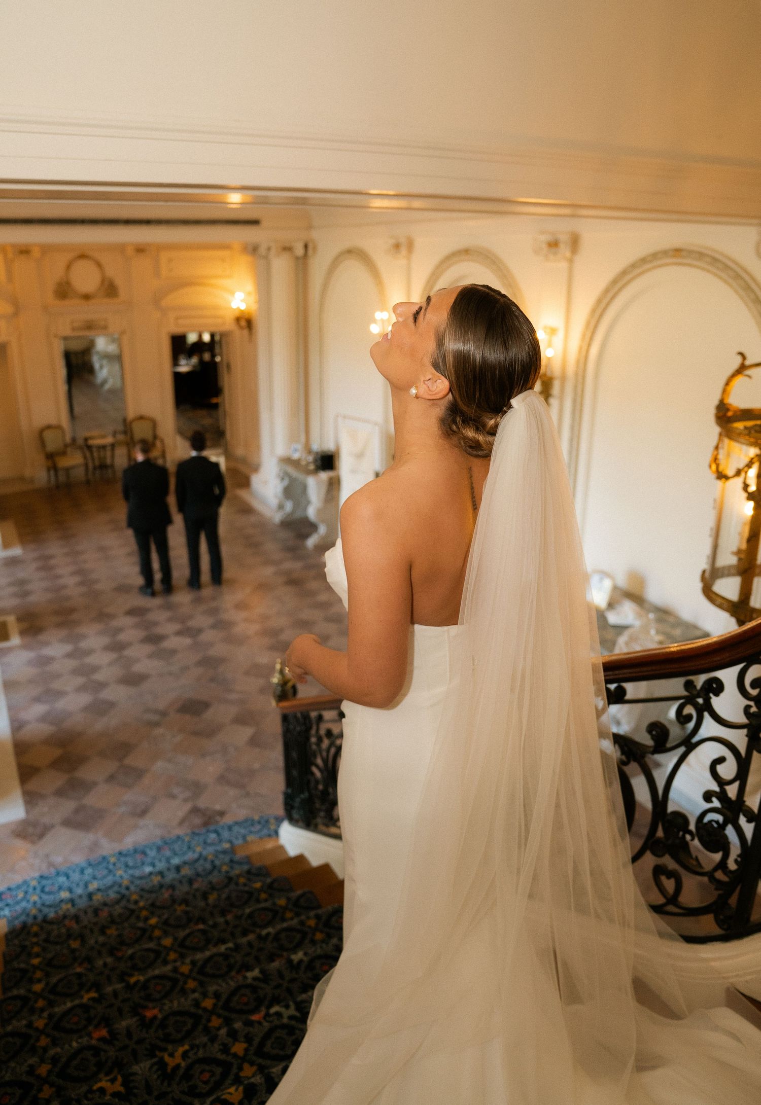 Bride in strapless white gown and veil stands on grand staircase of elegant venue, looking toward groom below.