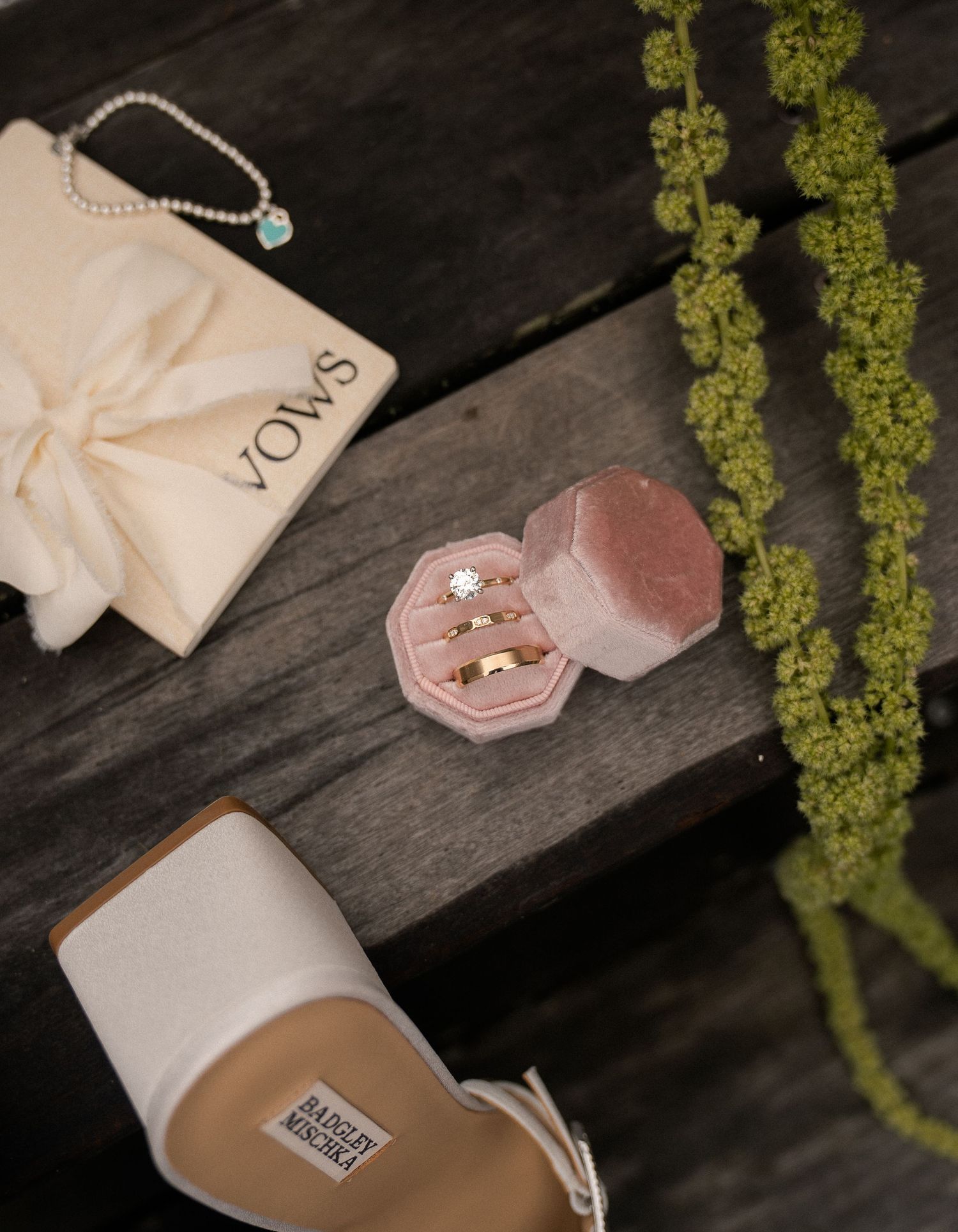 Pink velvet ring box with wedding bands, bridal shoes, pearl bracelet, and VOWS envelope on rustic wood surface.