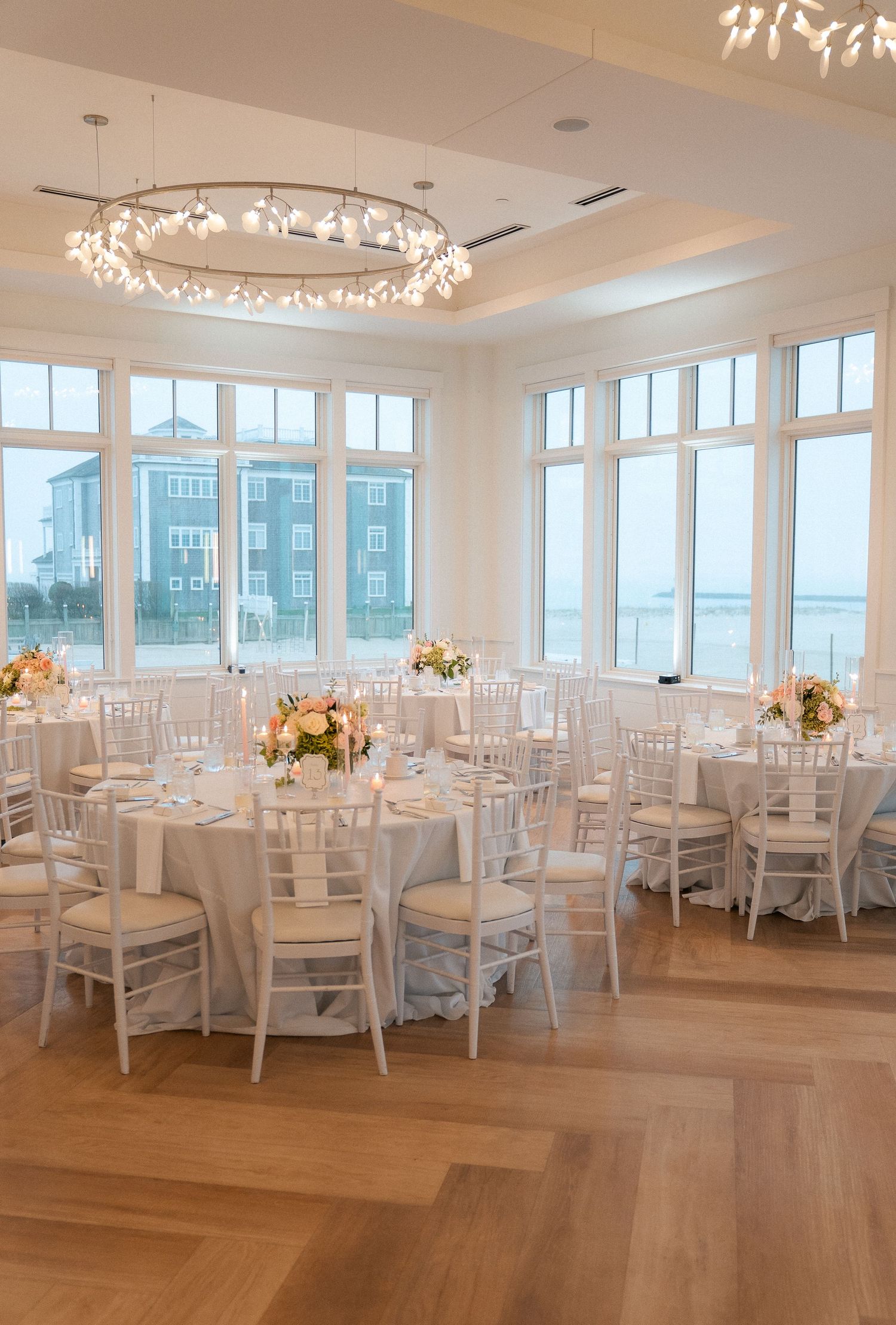 Elegant wedding reception hall with white chiavari chairs, pink floral centerpieces, and ocean views.