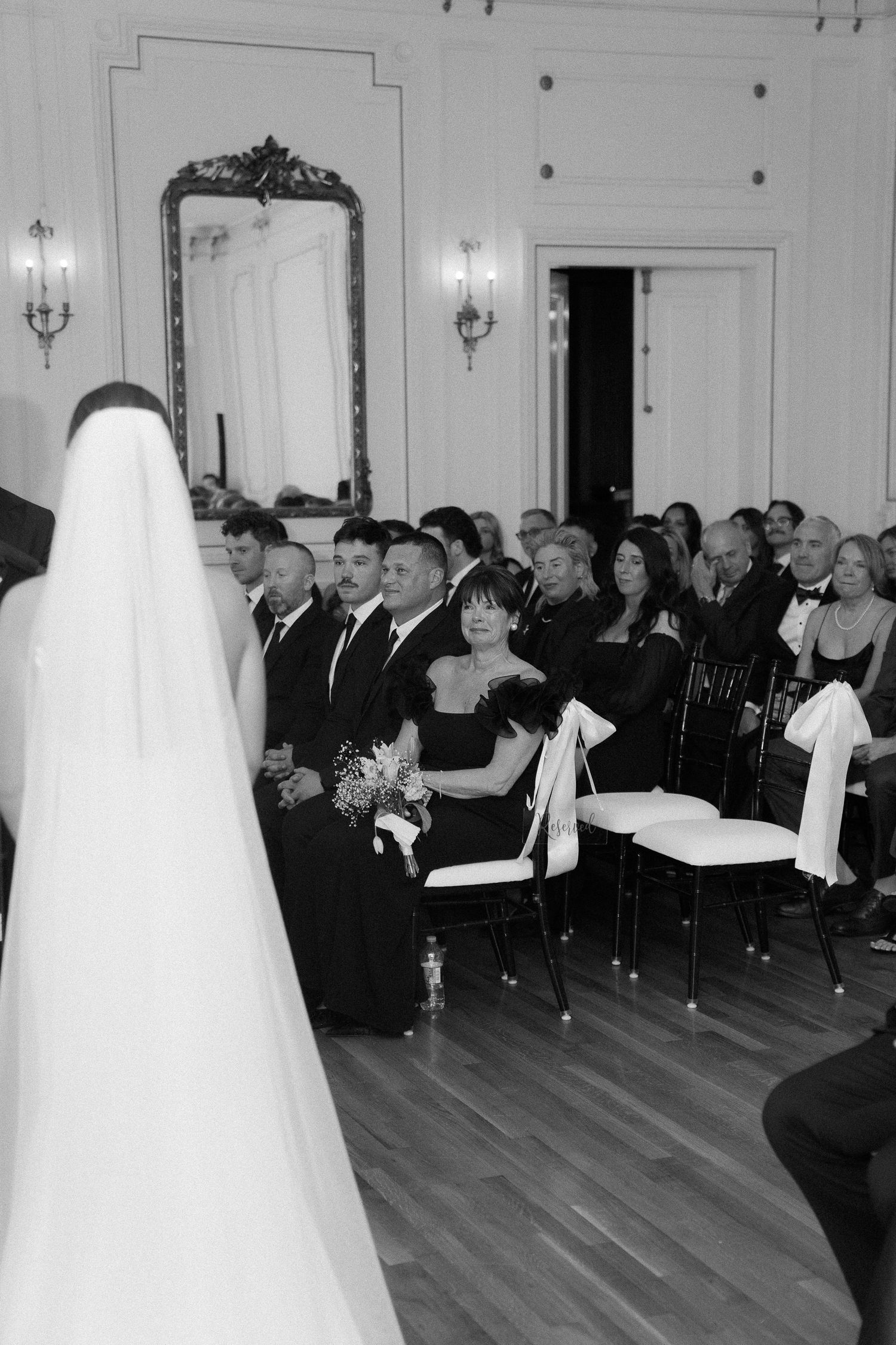 Black and white photo of a bride in veil walking down the aisle past seated wedding guests in an elegant ballroom.