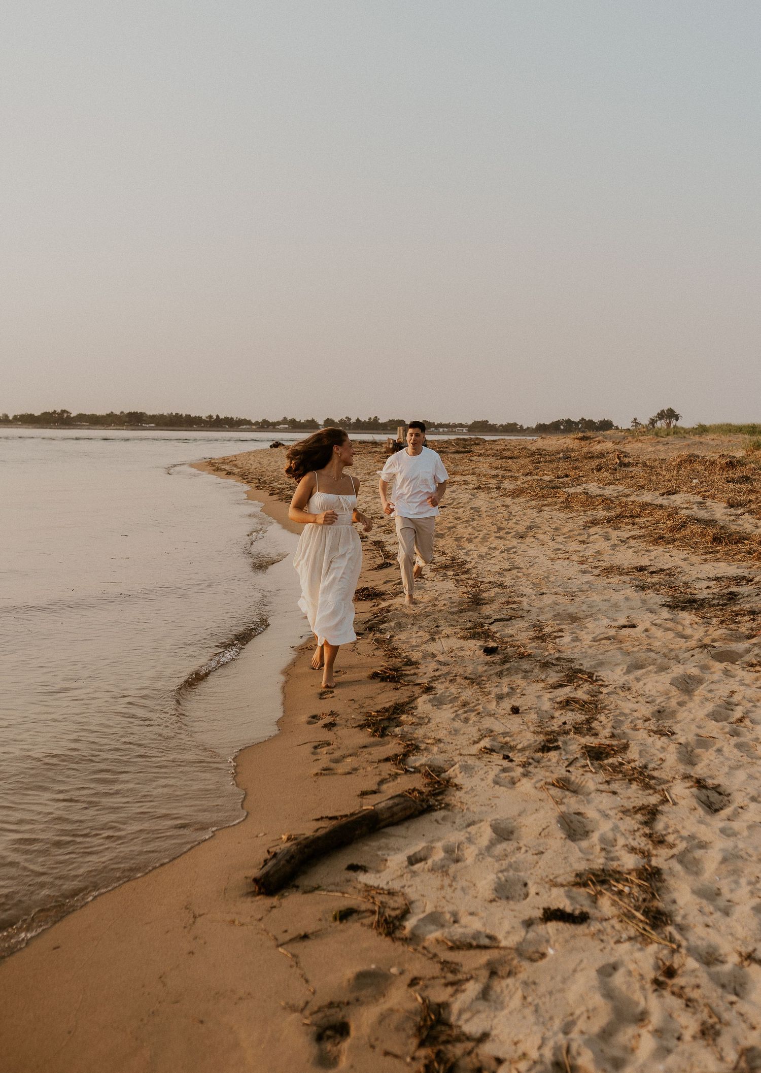 A couple in white clothing running playfully along a sandy beach at sunset.