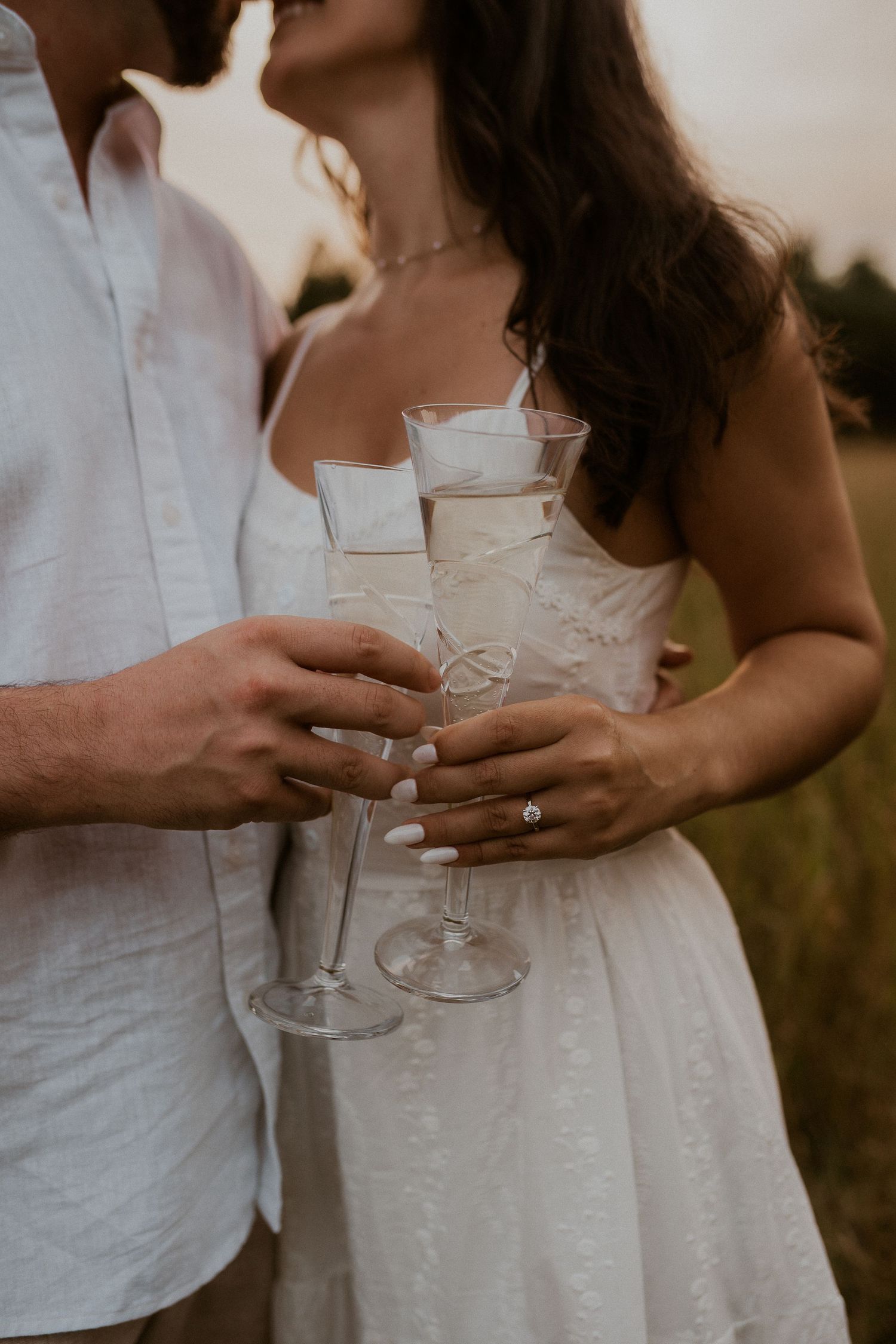 Couple holding champagne glasses together while wearing white outfits at sunset.