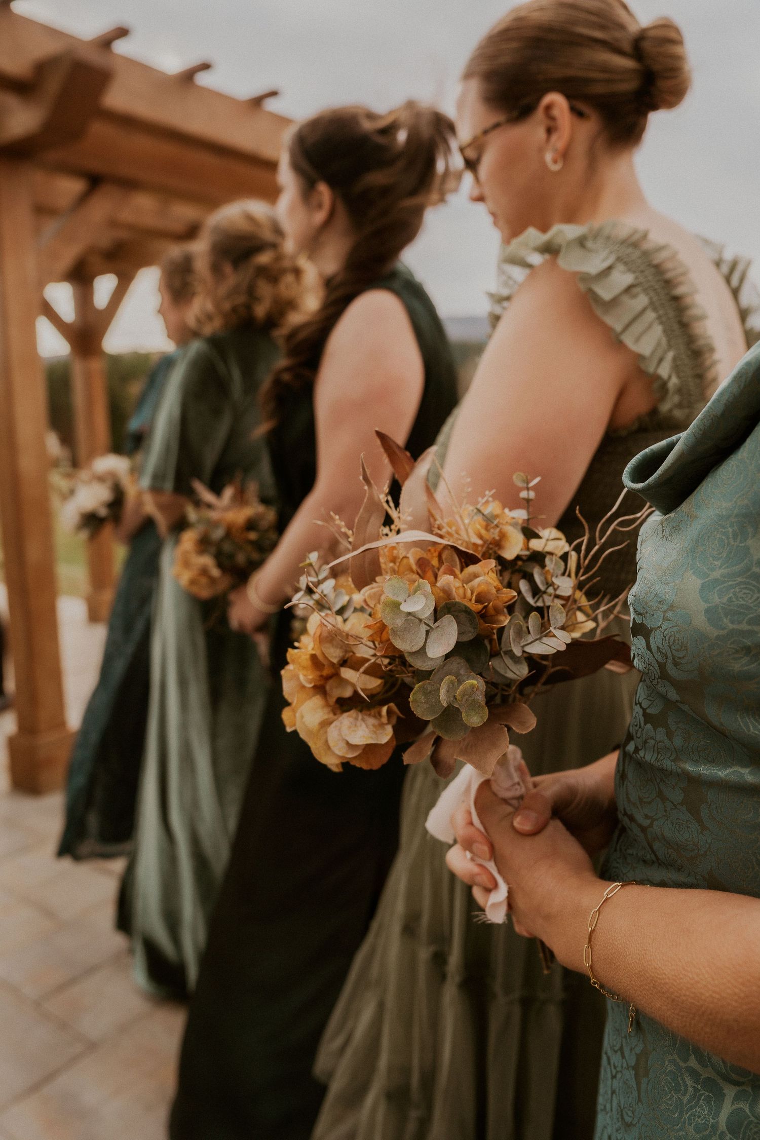Bridesmaids in sage green dresses hold autumn-colored bouquets at an outdoor wedding ceremony.