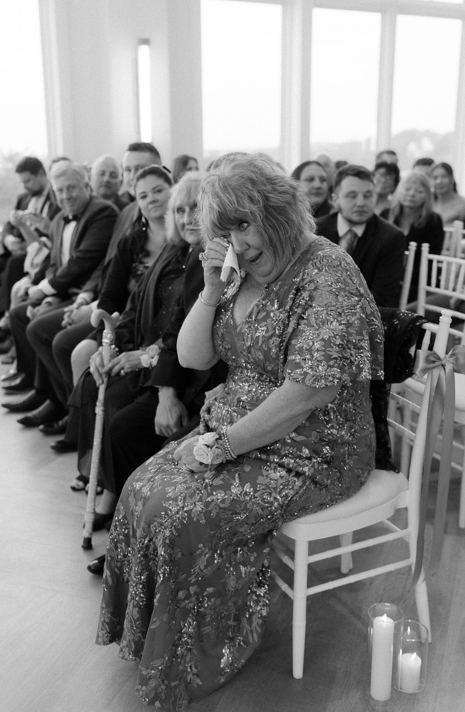 Emotional wedding guest wiping tears during ceremony seated among crowd in black and white photo.
