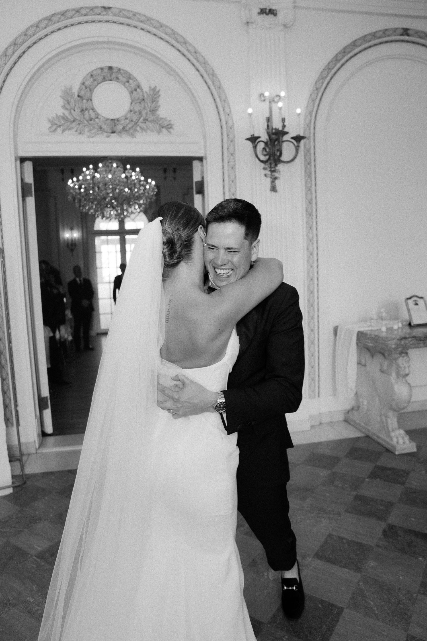 Black and white photo of groom laughing joyfully while embracing bride in ornate wedding venue hallway.