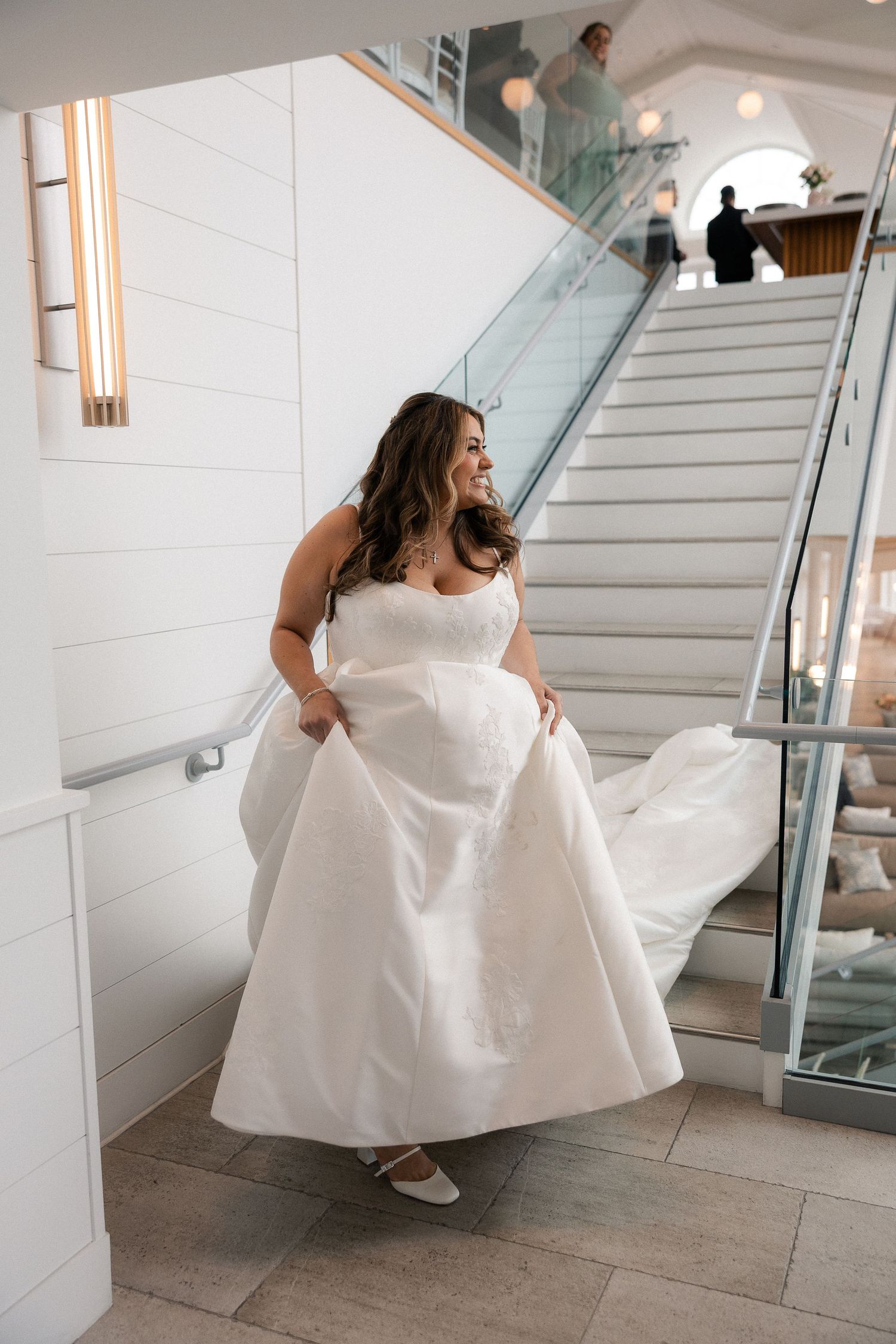 Bride in a strapless white ball gown lifts her skirt while smiling near a modern staircase.