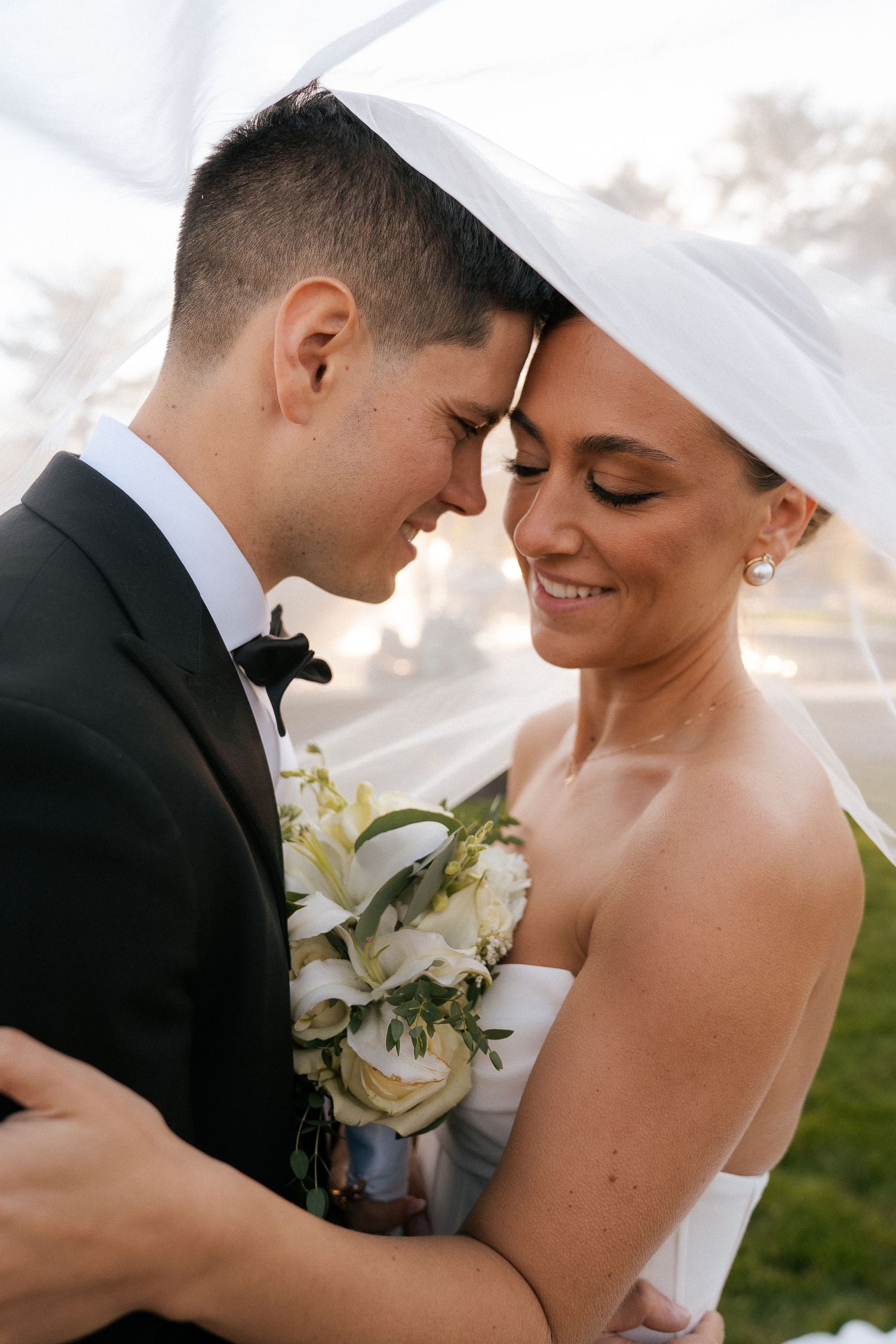 Bride and groom share an intimate forehead touch under a flowing veil, bride holding white floral bouquet.