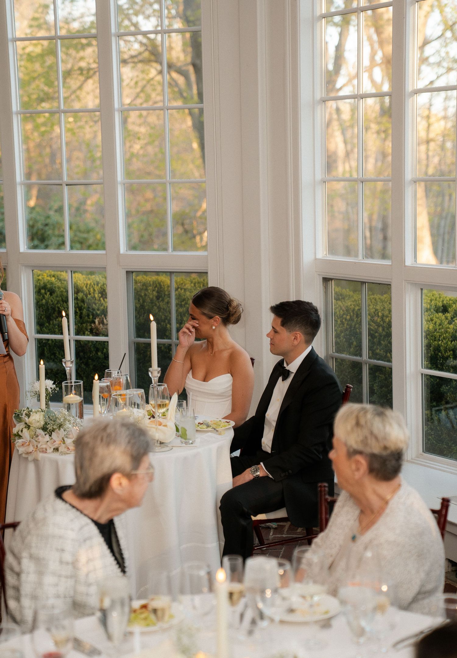 Bride and groom seated at sweetheart table during wedding reception in elegant sunlit venue with large windows.