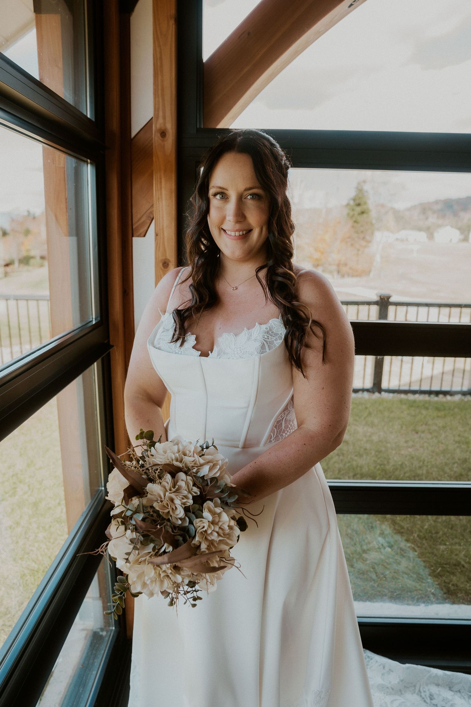 A bride in an elegant white gown smiles while holding a dried floral bouquet near large windows.