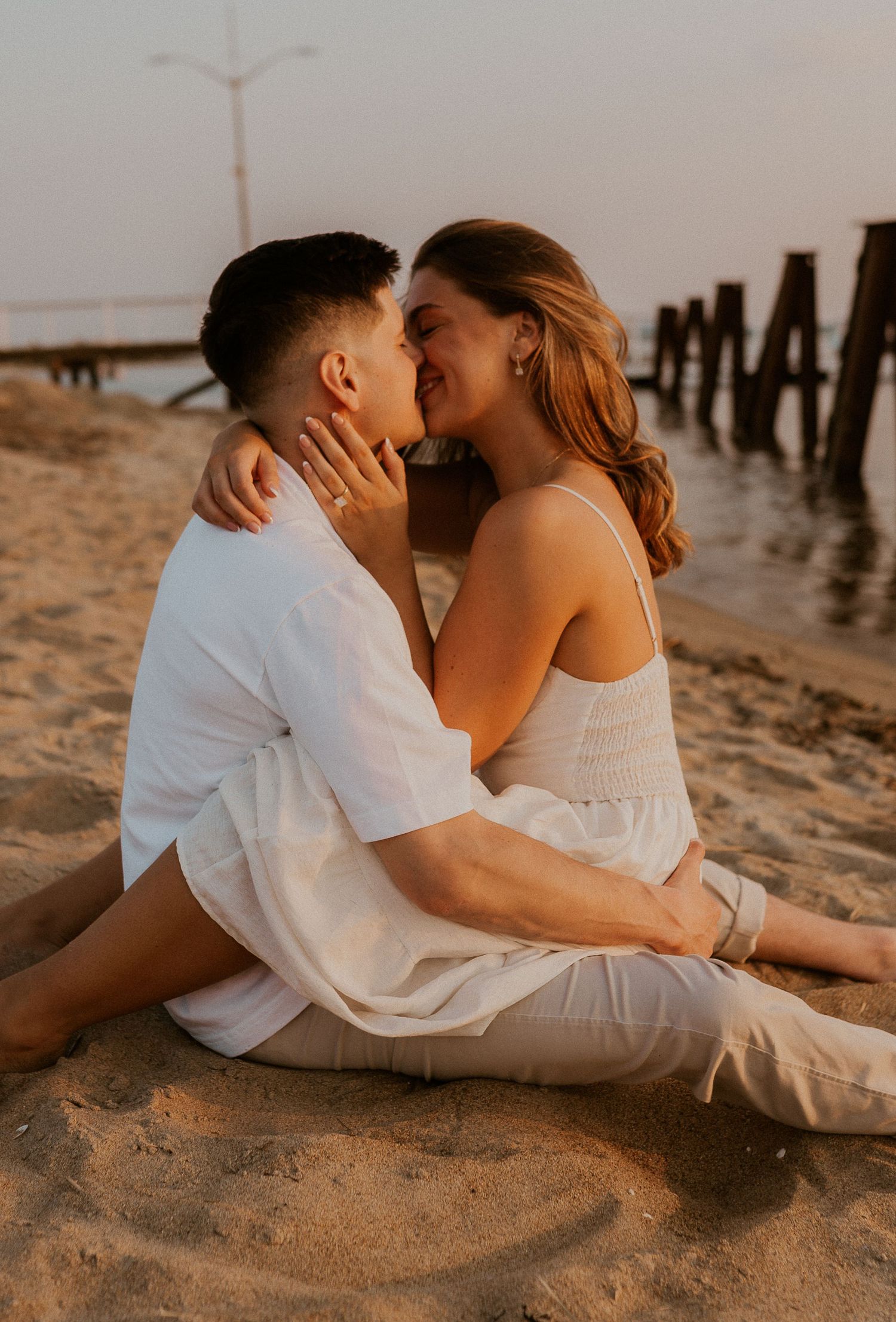 Romantic couple in white outfits embracing on sandy beach at sunset with wooden pier in background.