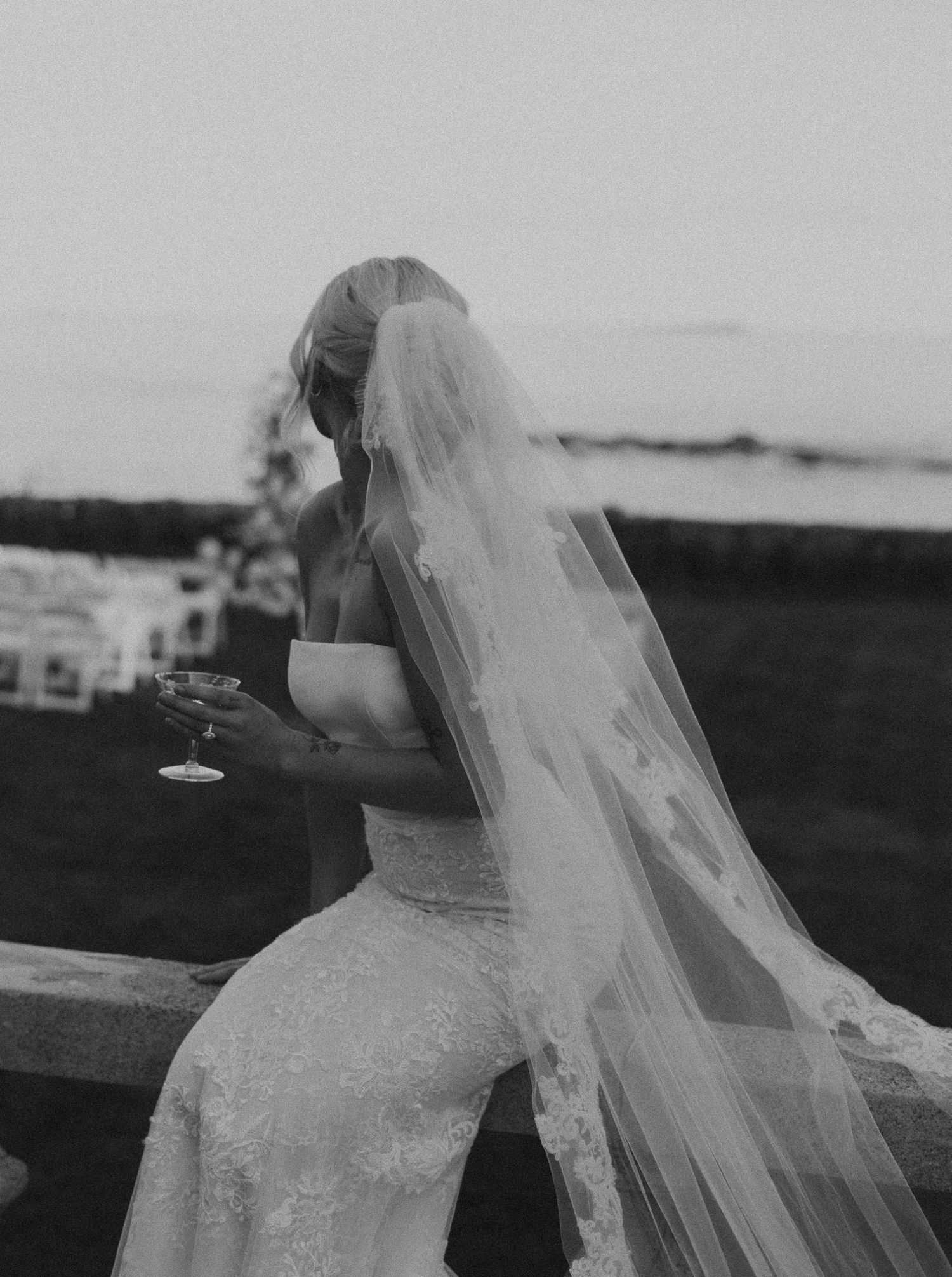 A bride in a flowing veil holds a drink while enjoying a scenic outdoor view.
