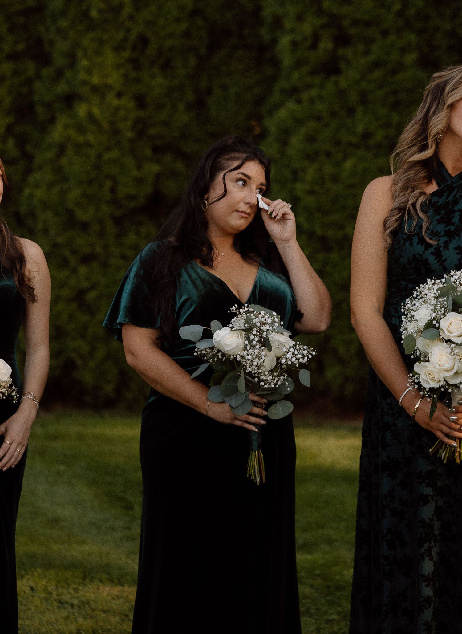 Bridesmaids in dark dresses holding white bouquets during outdoor wedding ceremony.